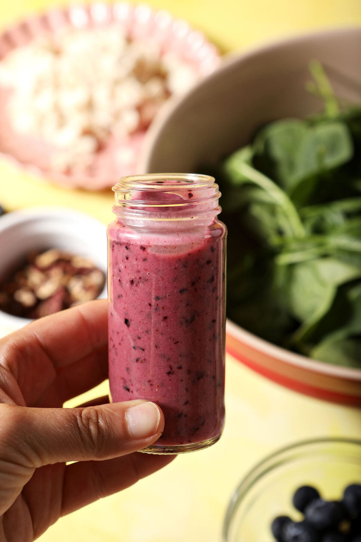 A hand holds a bottle of blueberry salad dressing above a yellow surface with salad ingredients on it
