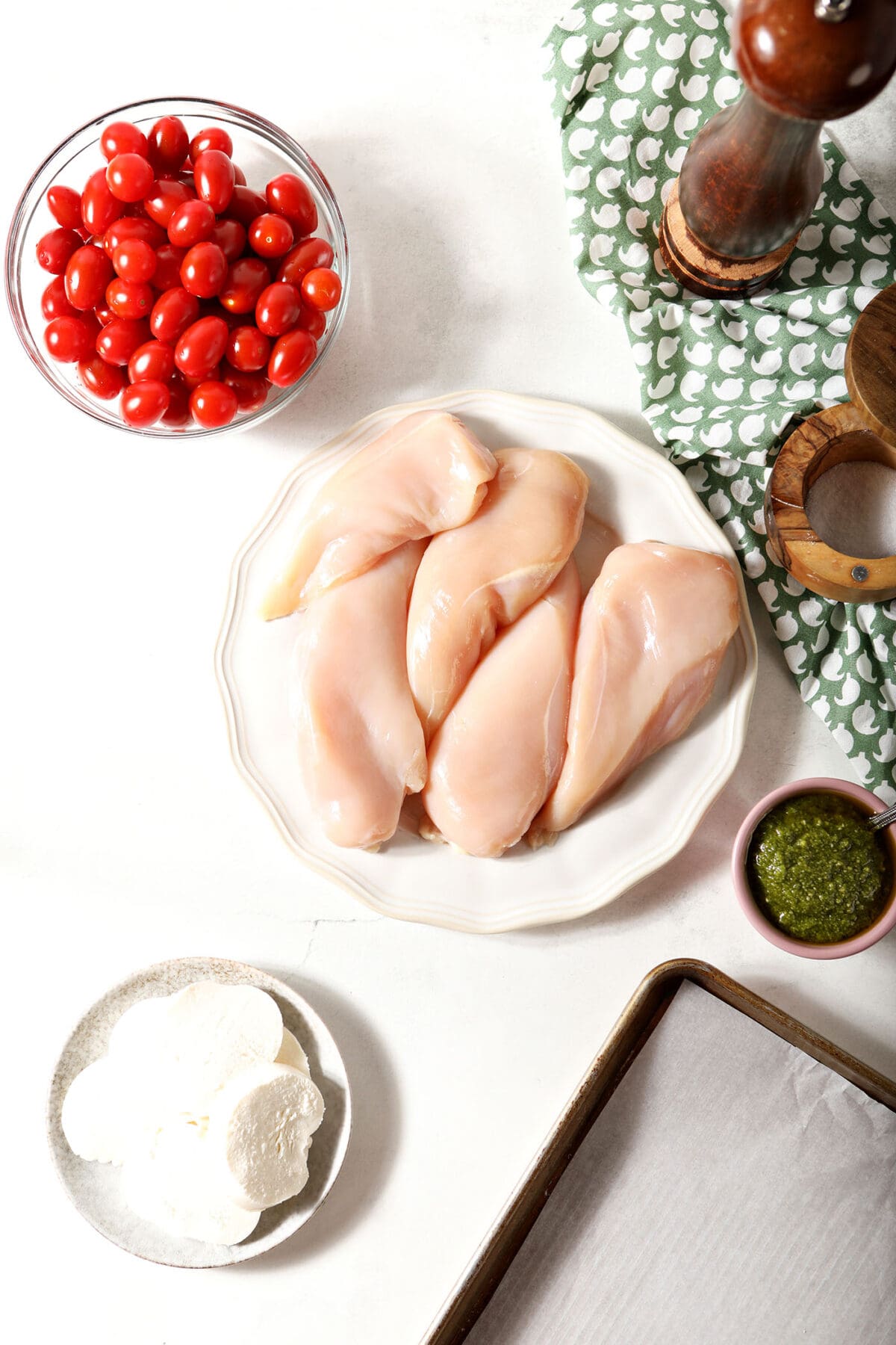 Ingredients to make baked caprese chicken on a white surface next to a green patterned linen