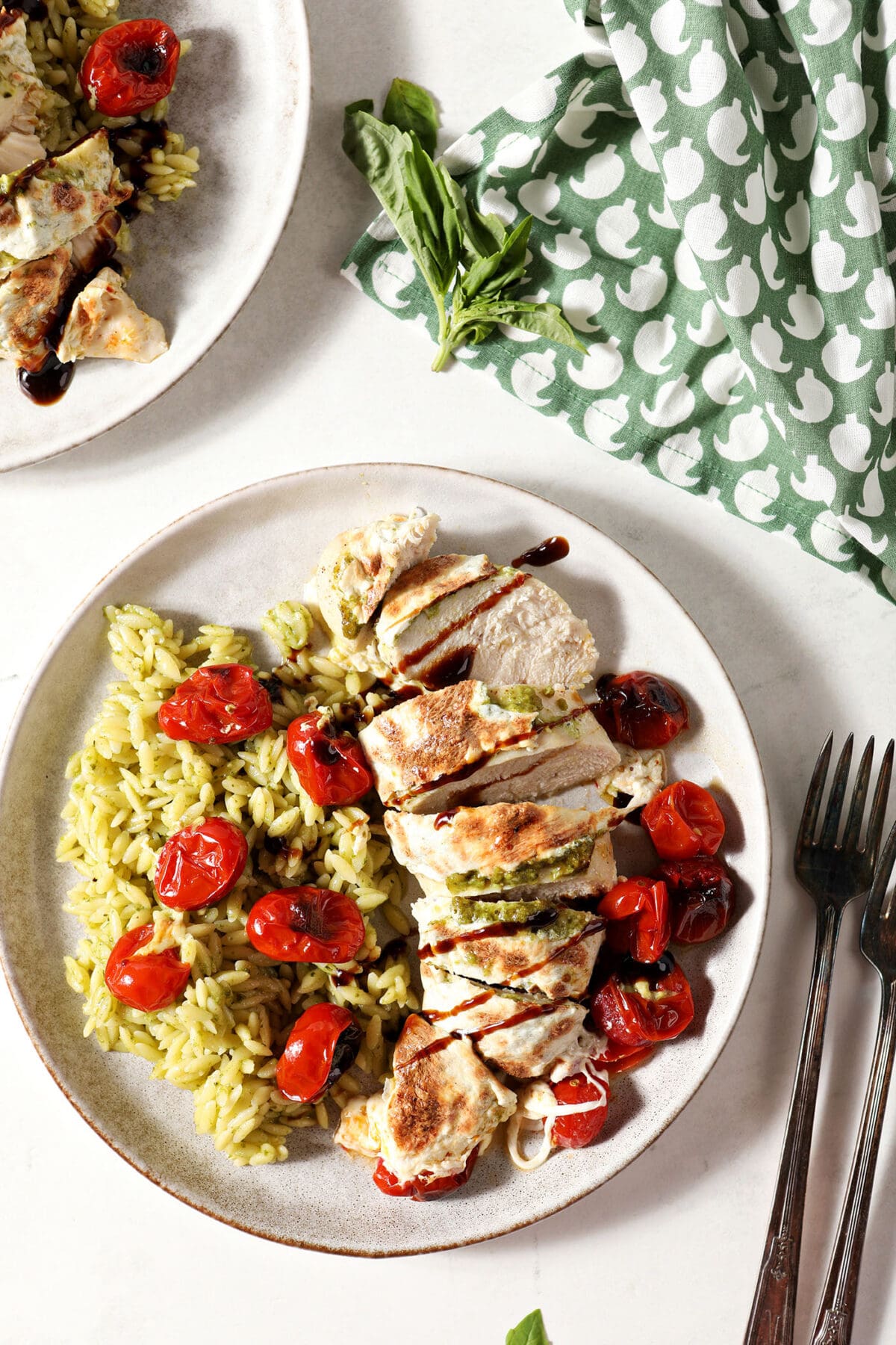 Two plates of baked caprese chicken on a white surface next to a green patterned linen