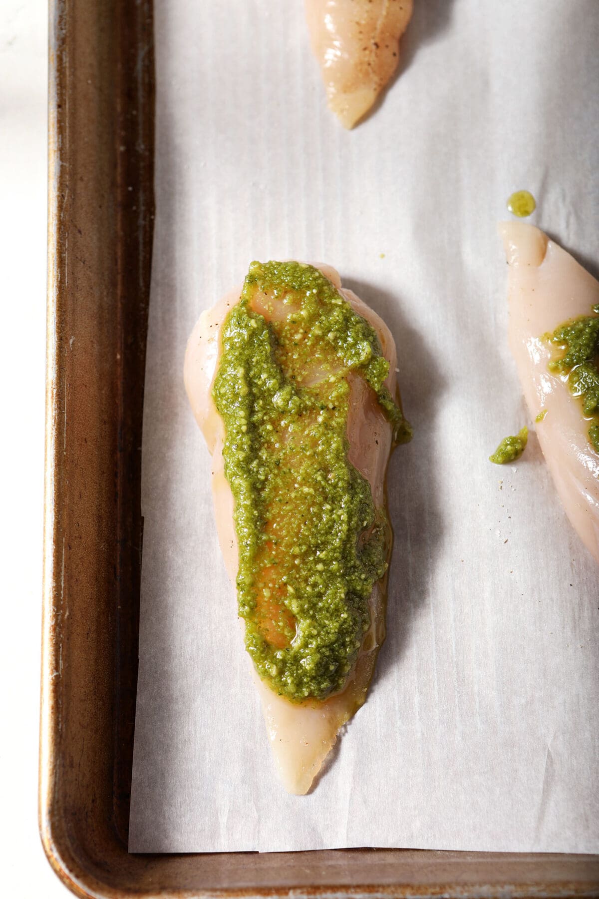 A chicken breast smeared with pesto on a parchment-lined sheet pan