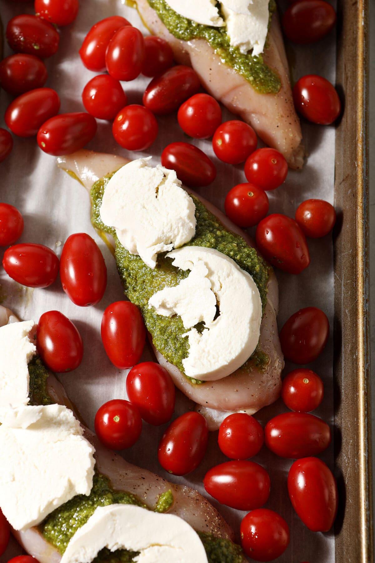 Raw chicken breasts topped with pesto and mozzarella surrounded by cherry tomatoes on a sheet pan