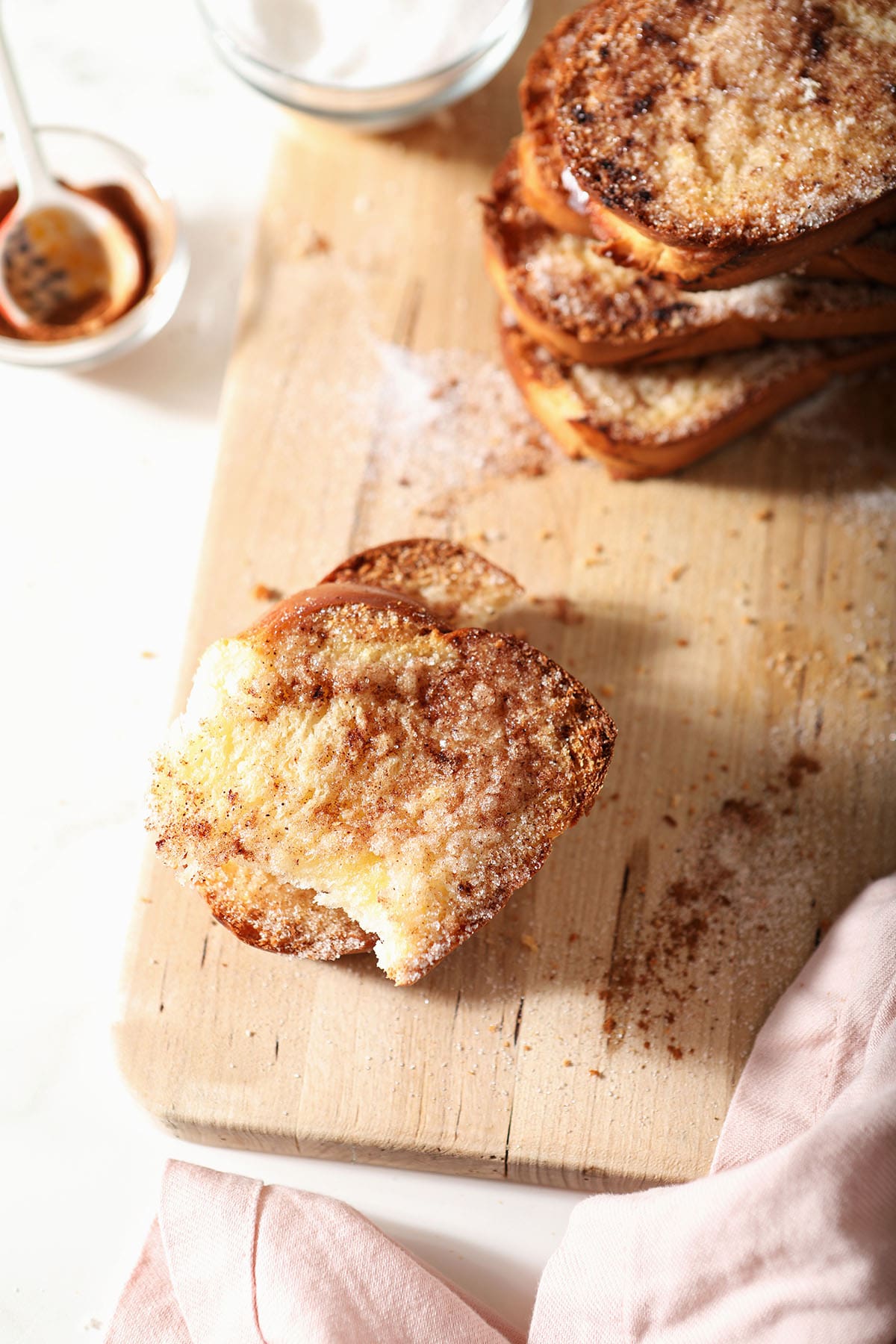 A bitten into piece of cinnamon sugar toast on a wooden cutting board with a pink linen and bowls of cinnamon and sugar