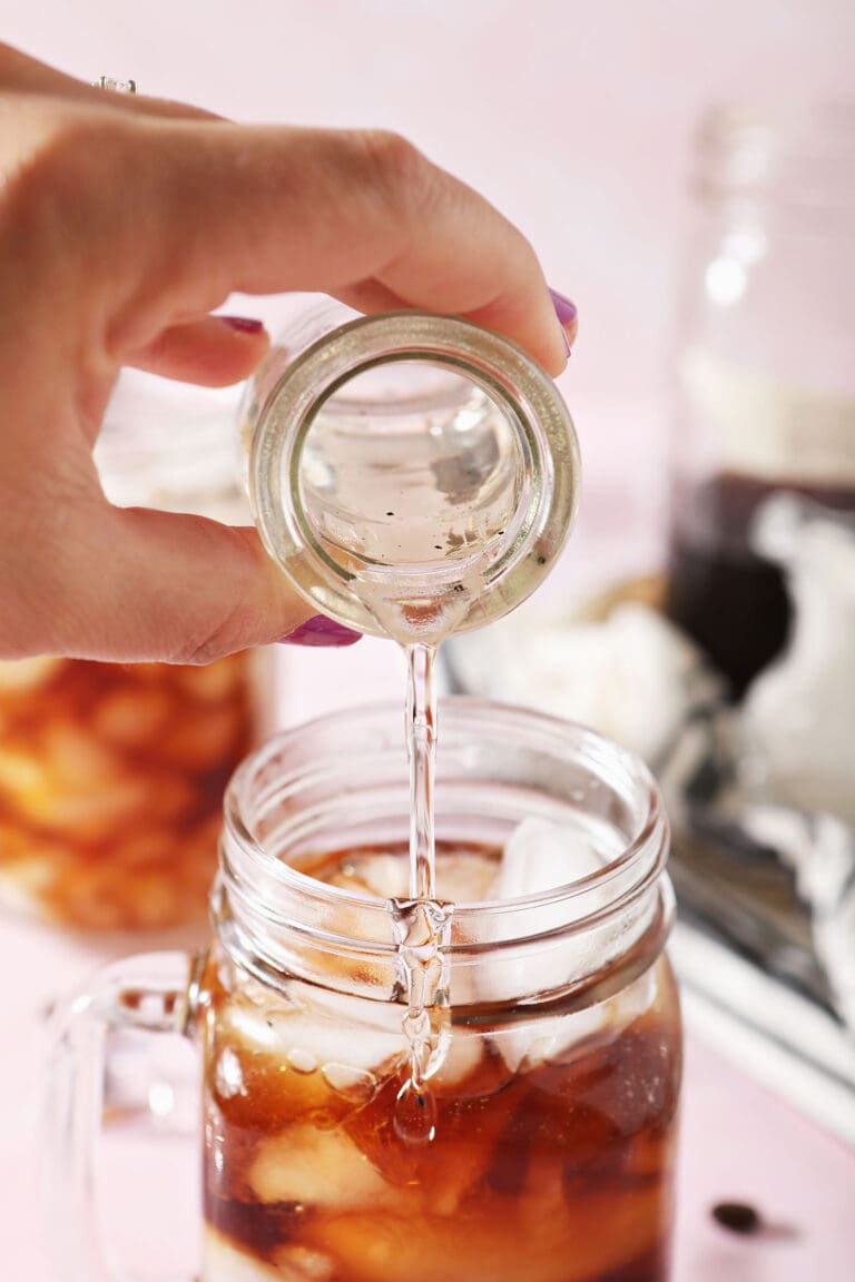 vanilla simple syrup pours into a glass of cold brew coffee on a pink surface