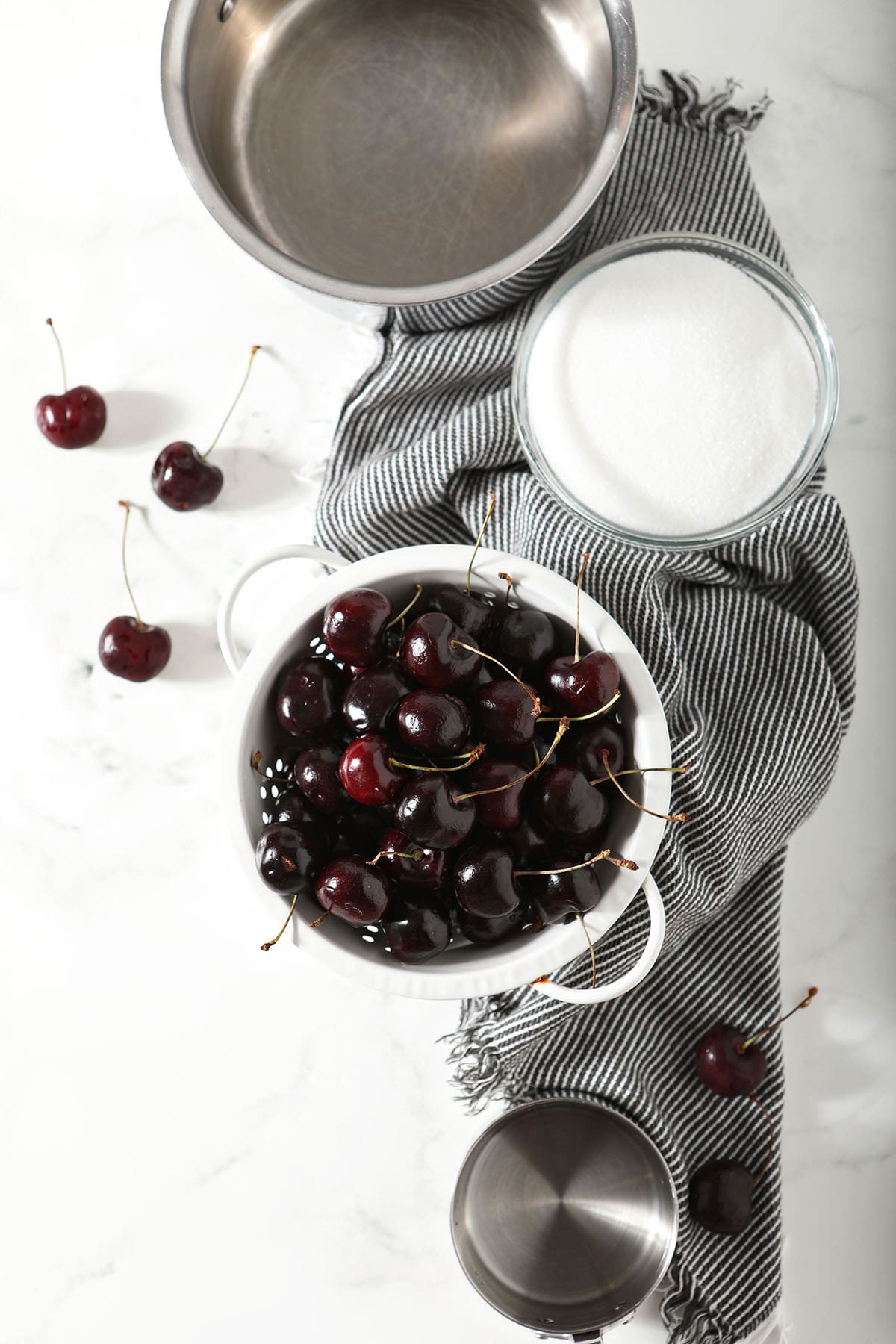 Fresh cherries in a colander next to a bowl of sugar and water showing ingredients to make cherry syrup