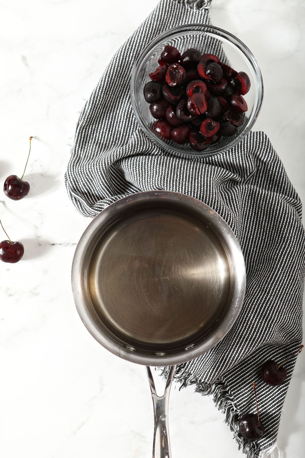 A saucepan of simple syrup next to a bowl of pitted cherries with a black and white striped linen