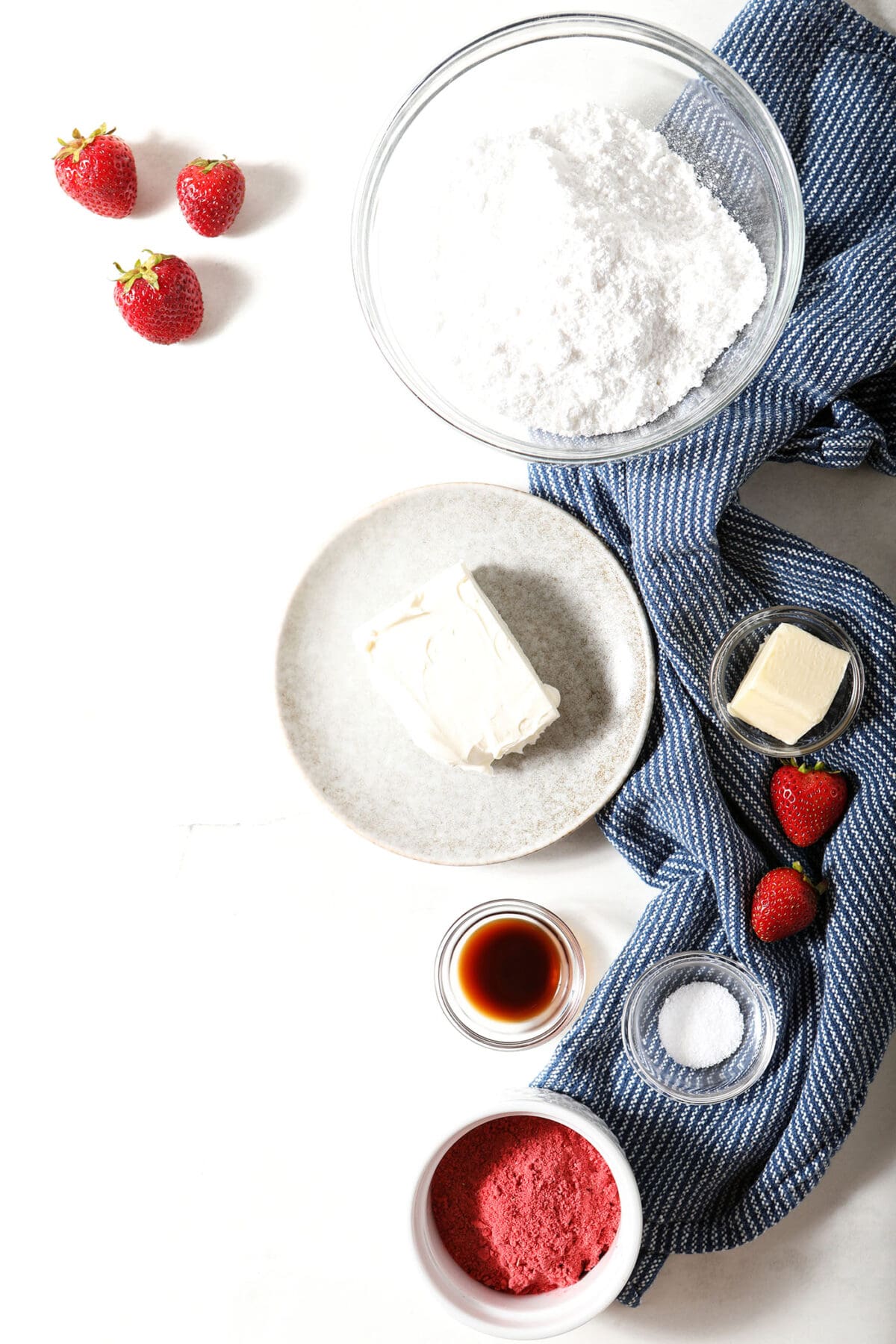 ingredients to make strawberry cream cheese frosting in bowls and plates on a stone surface with fresh berries