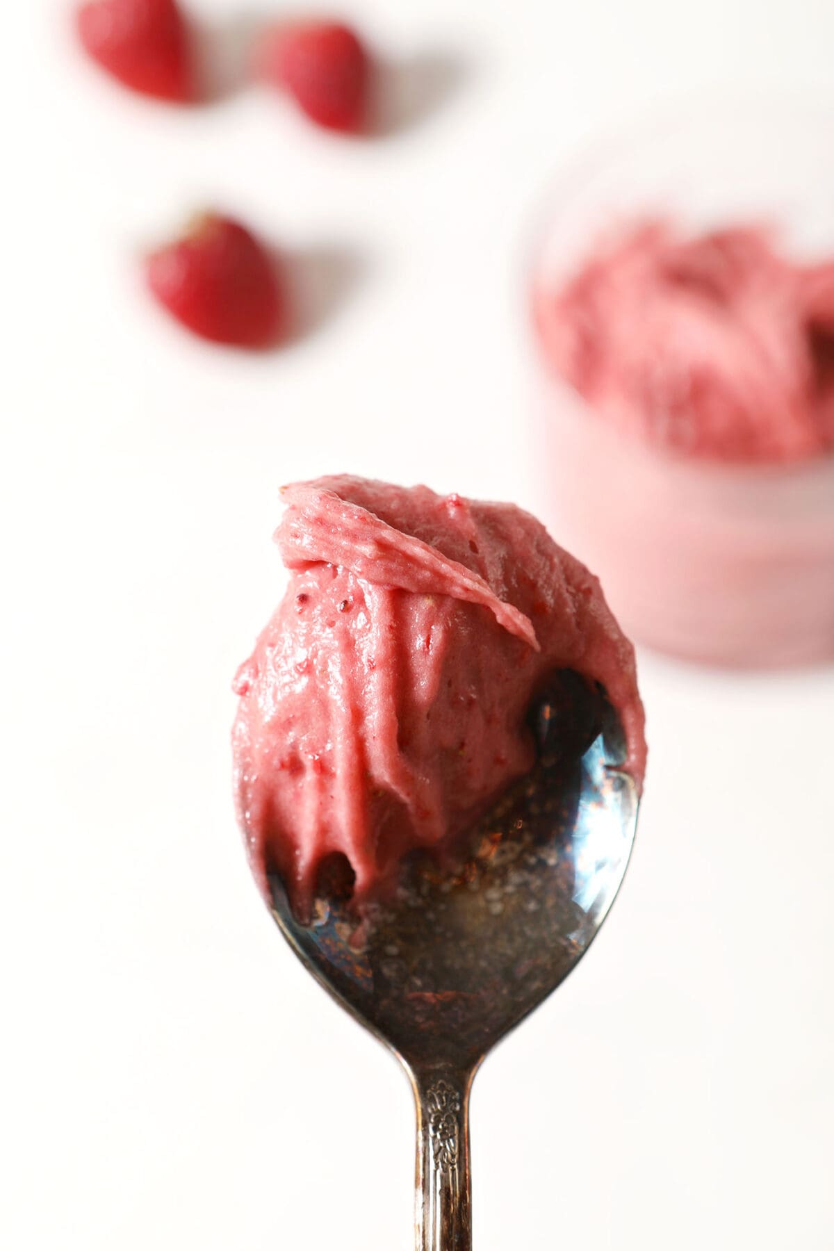 A spoonful of Strawberry Cream Cheese Frosting above a white stone surface with a bowl of frosting and strawberries