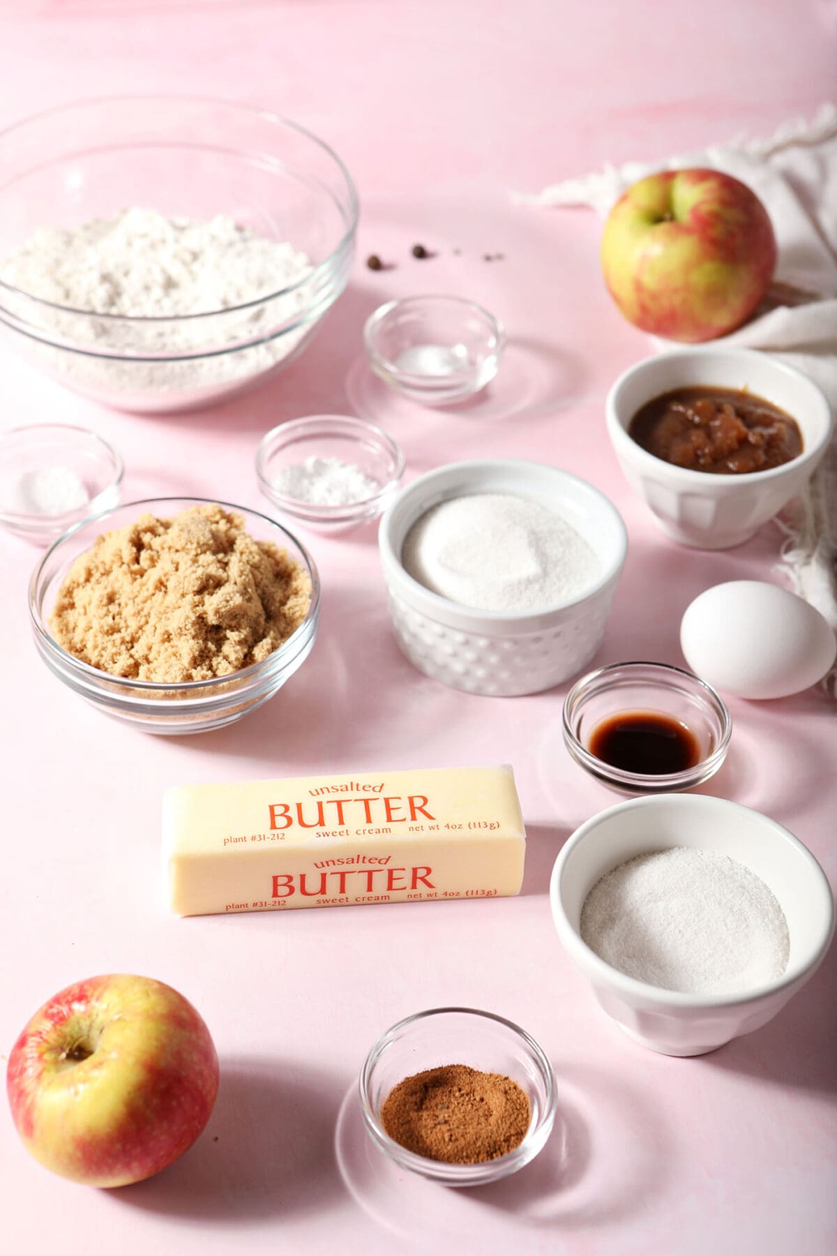Ingredients to make apple snickerdoodles in bowls surrounded by spices and apples on a pink surface