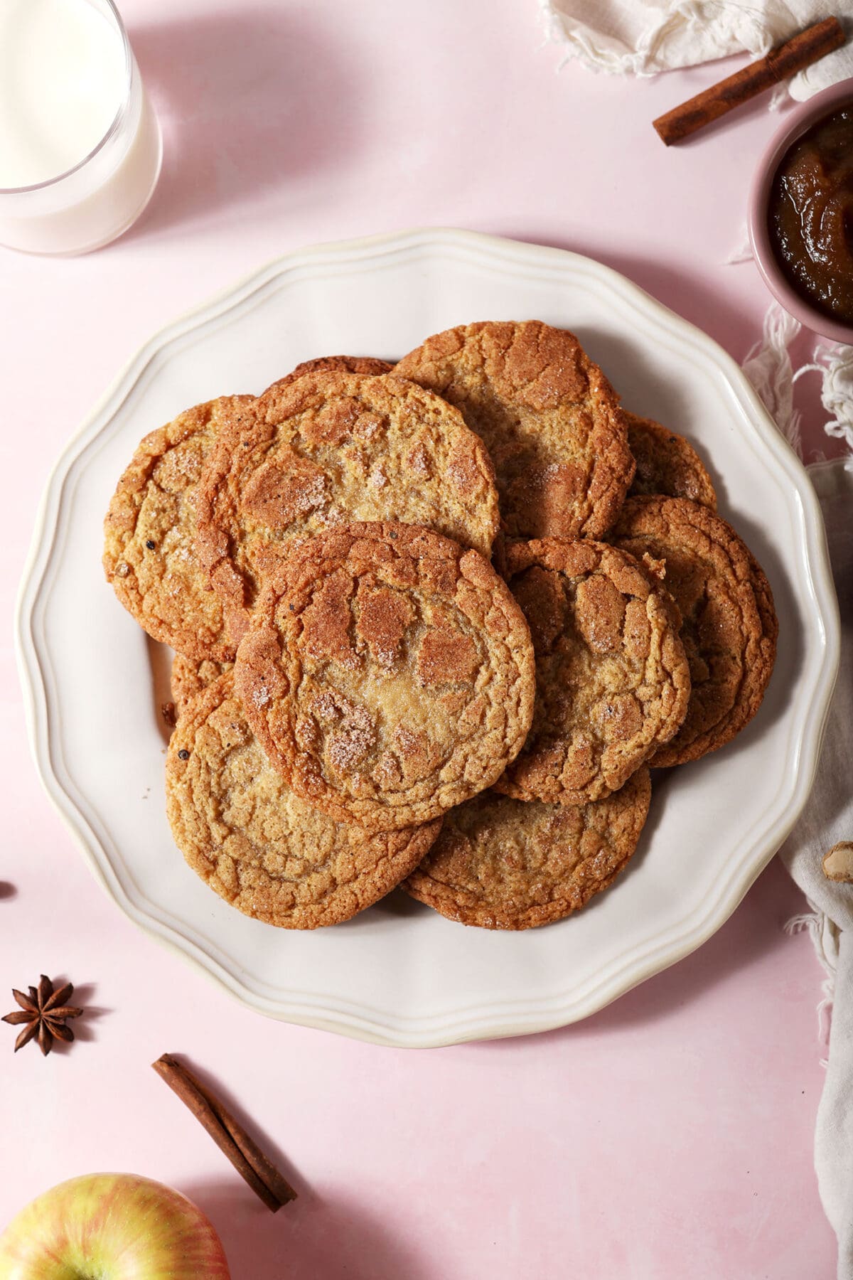 A plate of stacked apple doodle cookies on a pink surface next to warming spices and a glass of milk