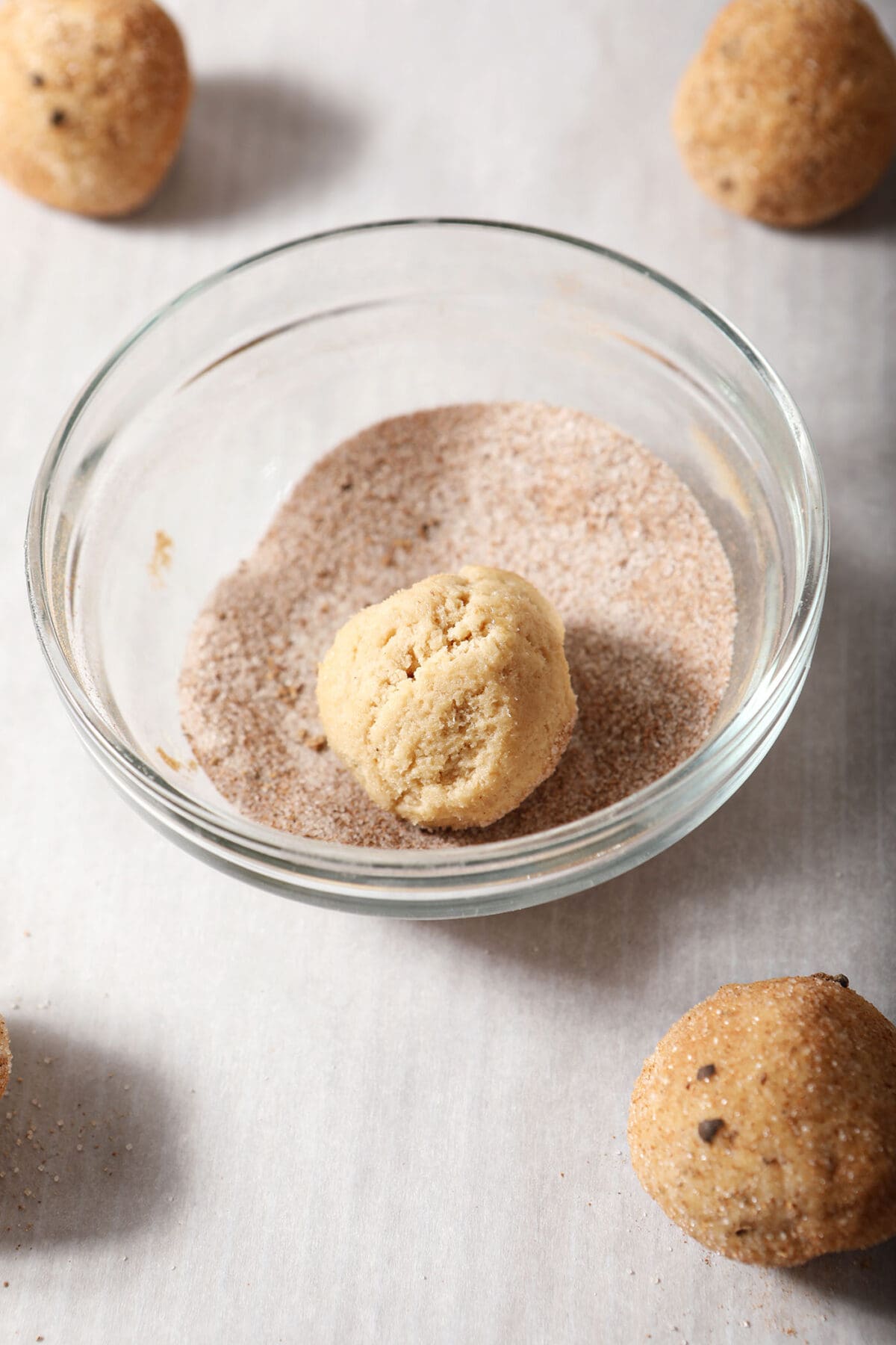 A cookie dough round in a bowl of sugar and spices sits on a sheet pan between other cookie dough balls