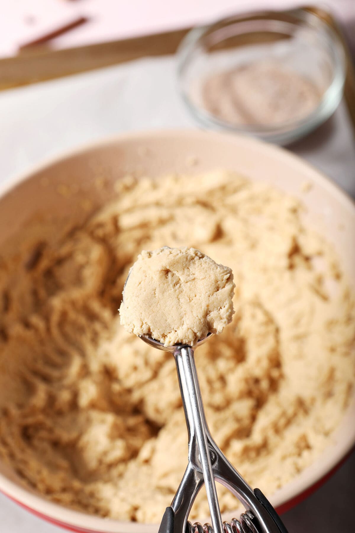 A cookie scoop holds a portion of cookie dough above a bowl and a lined sheet pan