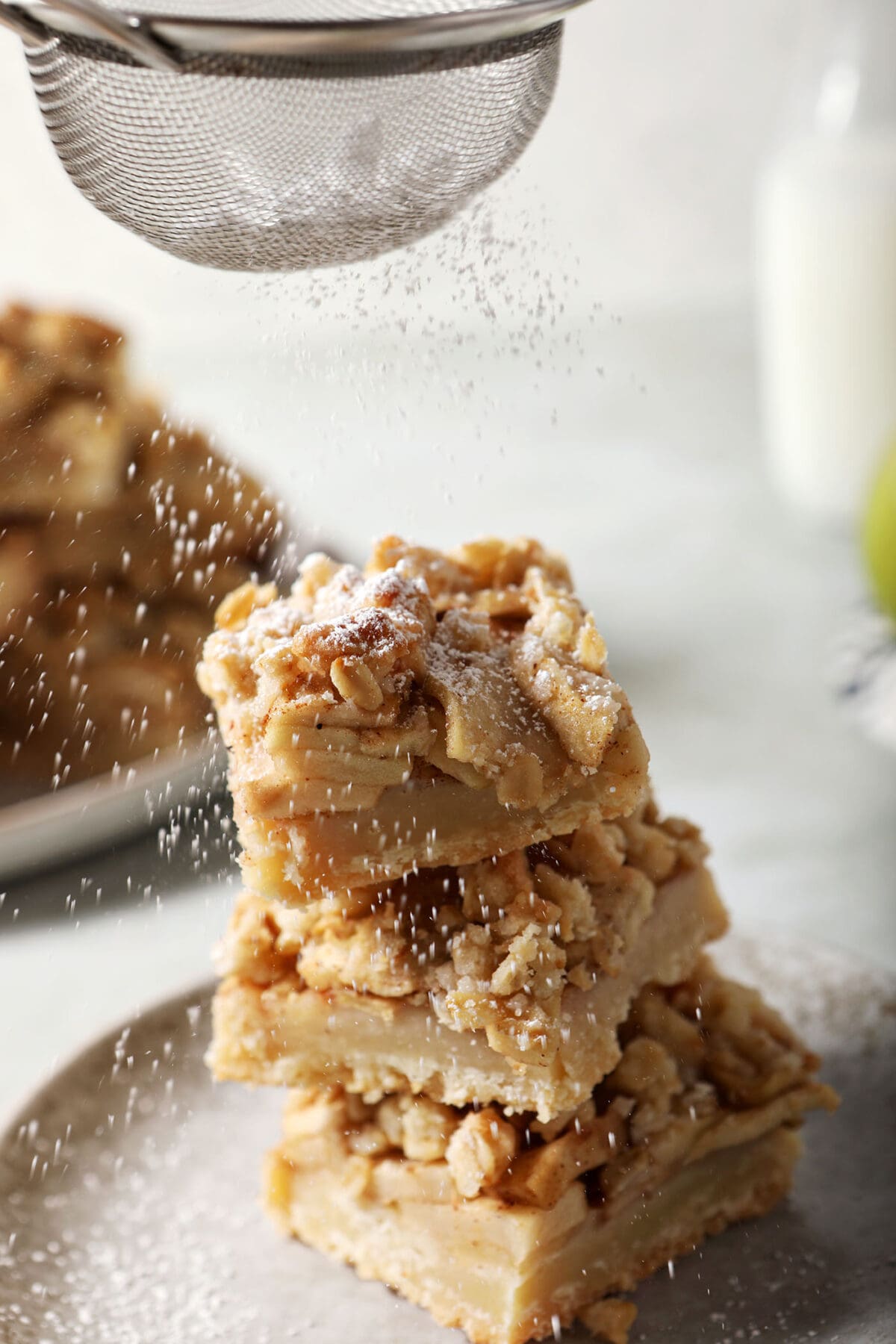 Powdered sugar sprinkles on top of a stack of apple pie bars on a plate