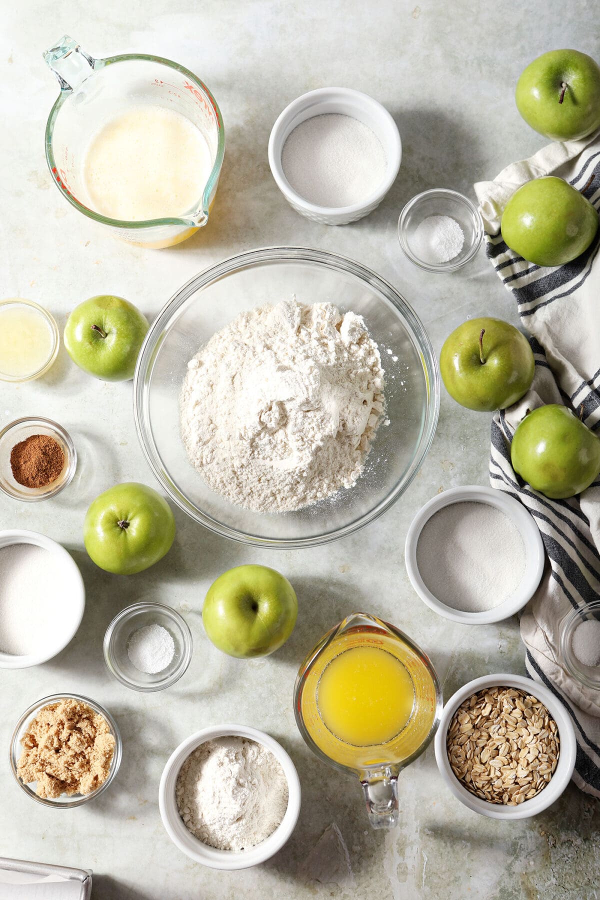 Ingredients to make homemade apple pie bars in bowls on a green surface next to a black and white linen