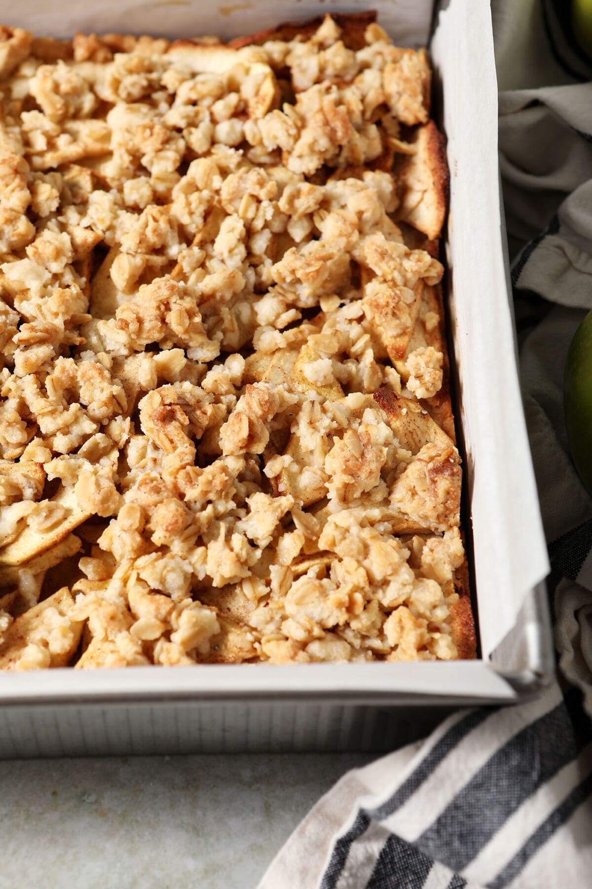 Baked apple pie bars in a square baking dish on a green surface next to a black and white striped linen