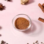 a bowl of homemade apple pie spice on a pink surface surrounded by whole spices