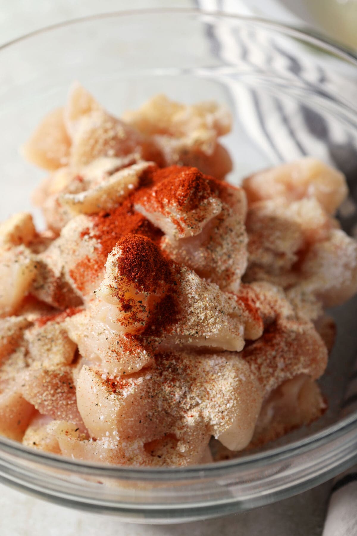 Close up of seasonings on top of chopped chicken breasts in a glass bowl on an olive green surface