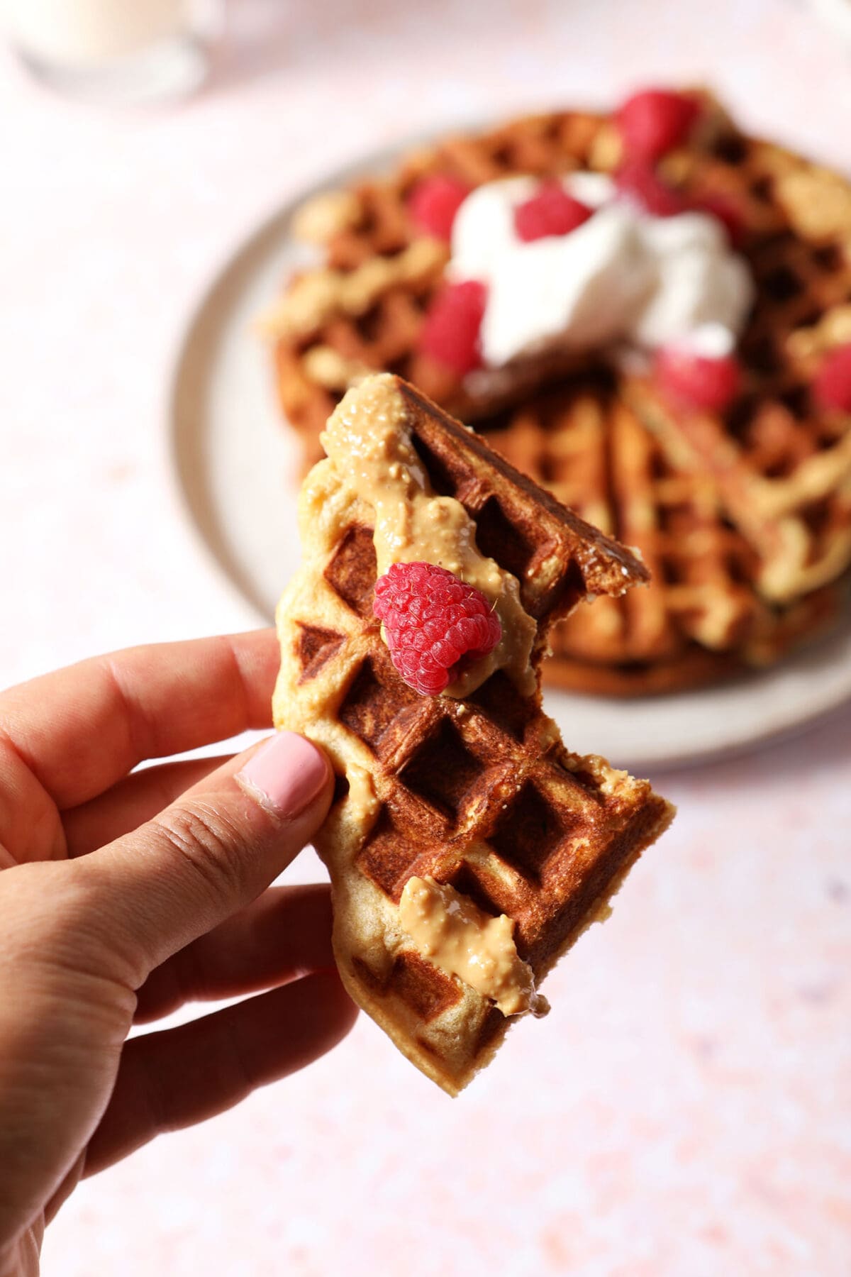 A hand holds a bitten-into waffle topped with peanut butter and raspberry above a plate of waffles