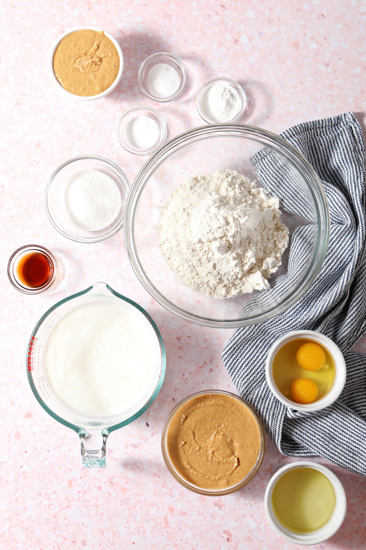 Ingredients to make Peanut Butter Waffles in bowls on a pink surface with a blue striped towel