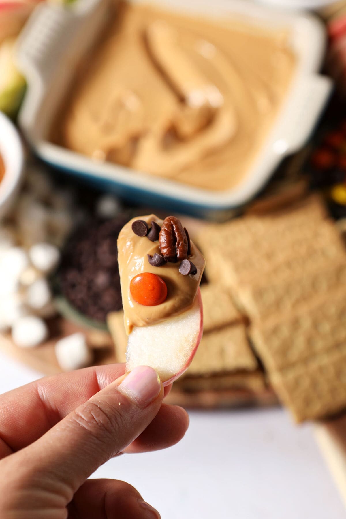 A hand holds an apple slice coated in caramel dip, chocolate chips, pecans and Reese’s pieces