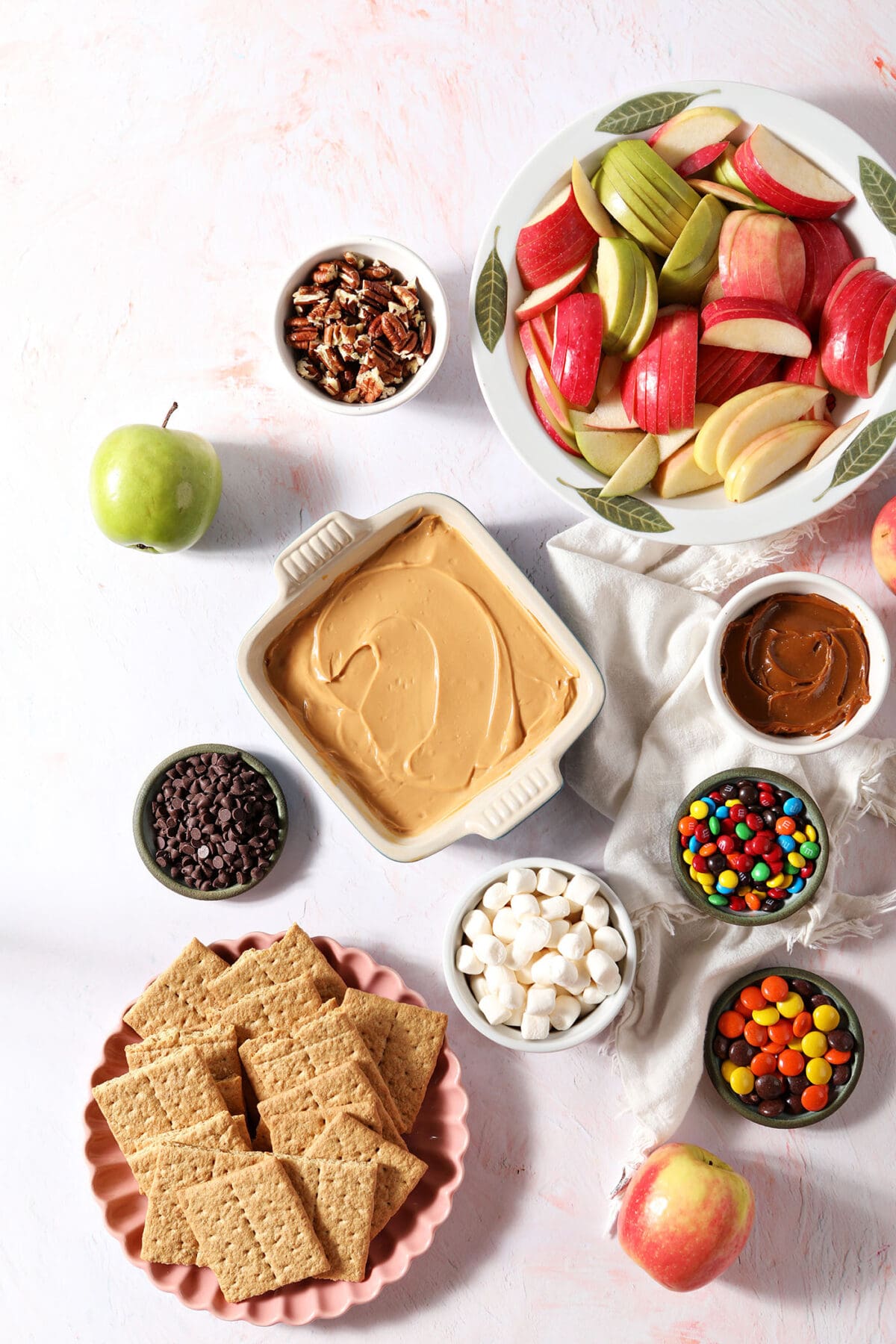 Ingredients to make a Caramel Apple “Charcuterie” Board in bowls on a light pink surface