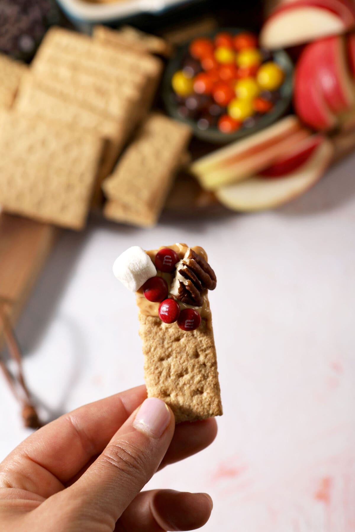 A hand holds a graham cracker coated in caramel dip and other toppings from a caramel apple board