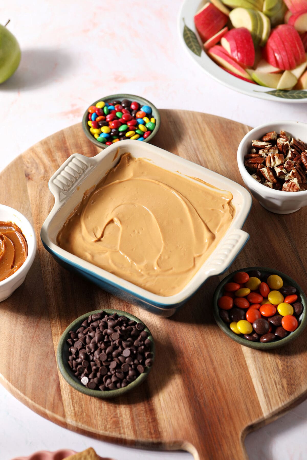 Bowls of candies and other ingredients around a container of caramel dip on a wooden board