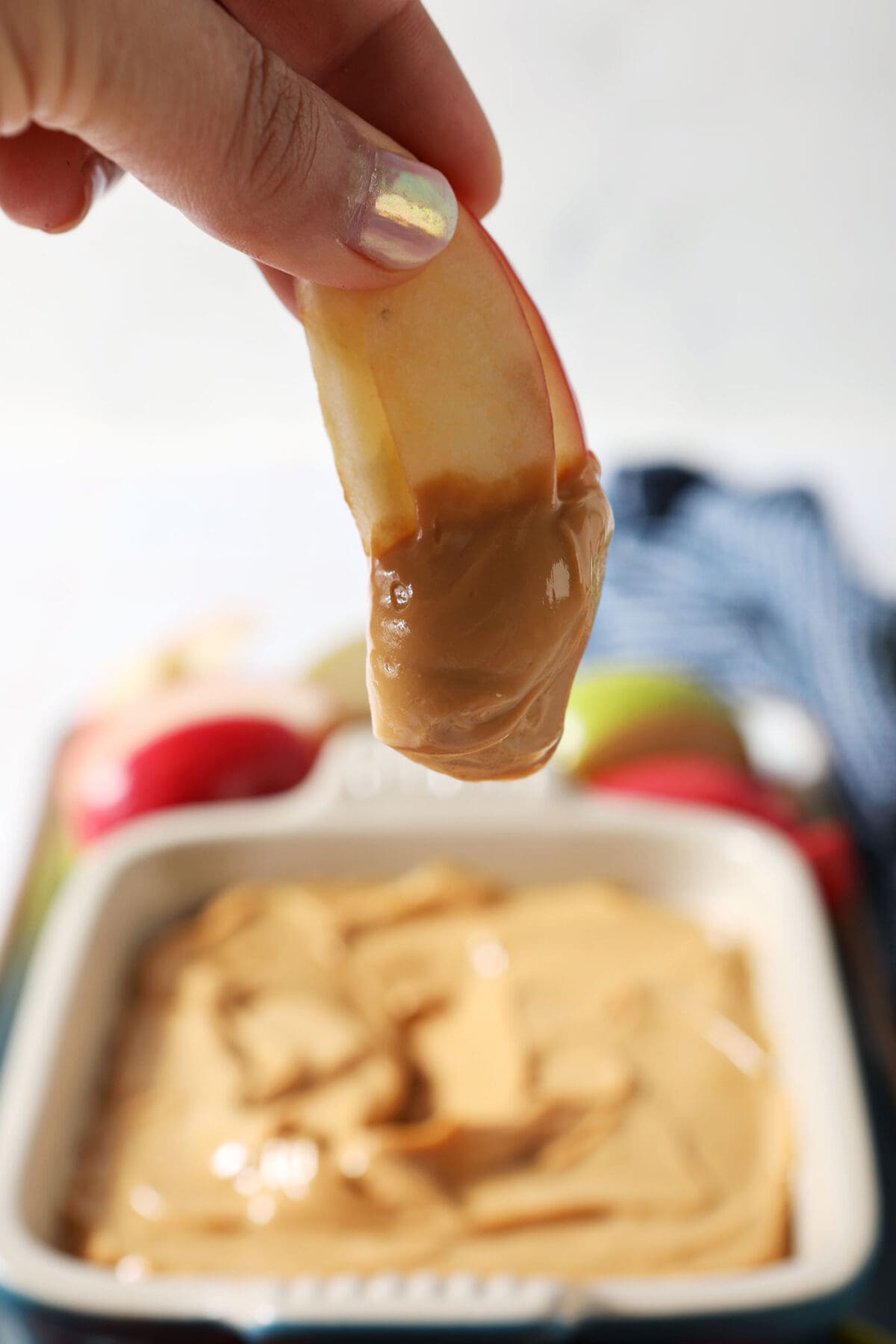 A hand holds an apple slice dipped in Dulce de Leche Caramel Dip above a platter