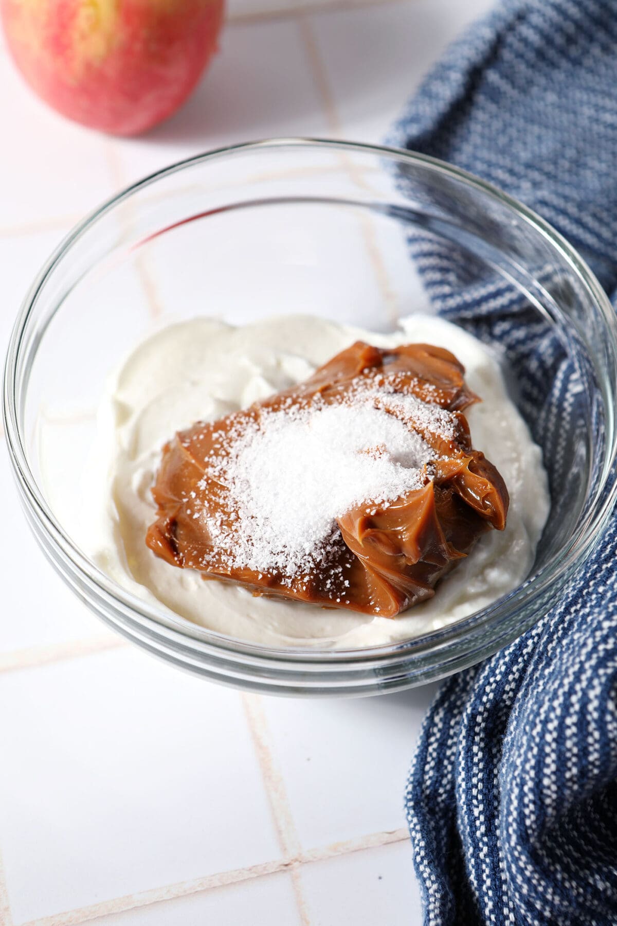 Greek yogurt, dulce de leche and salt in a mixing bowl on a white tiled surface next to a blue linen