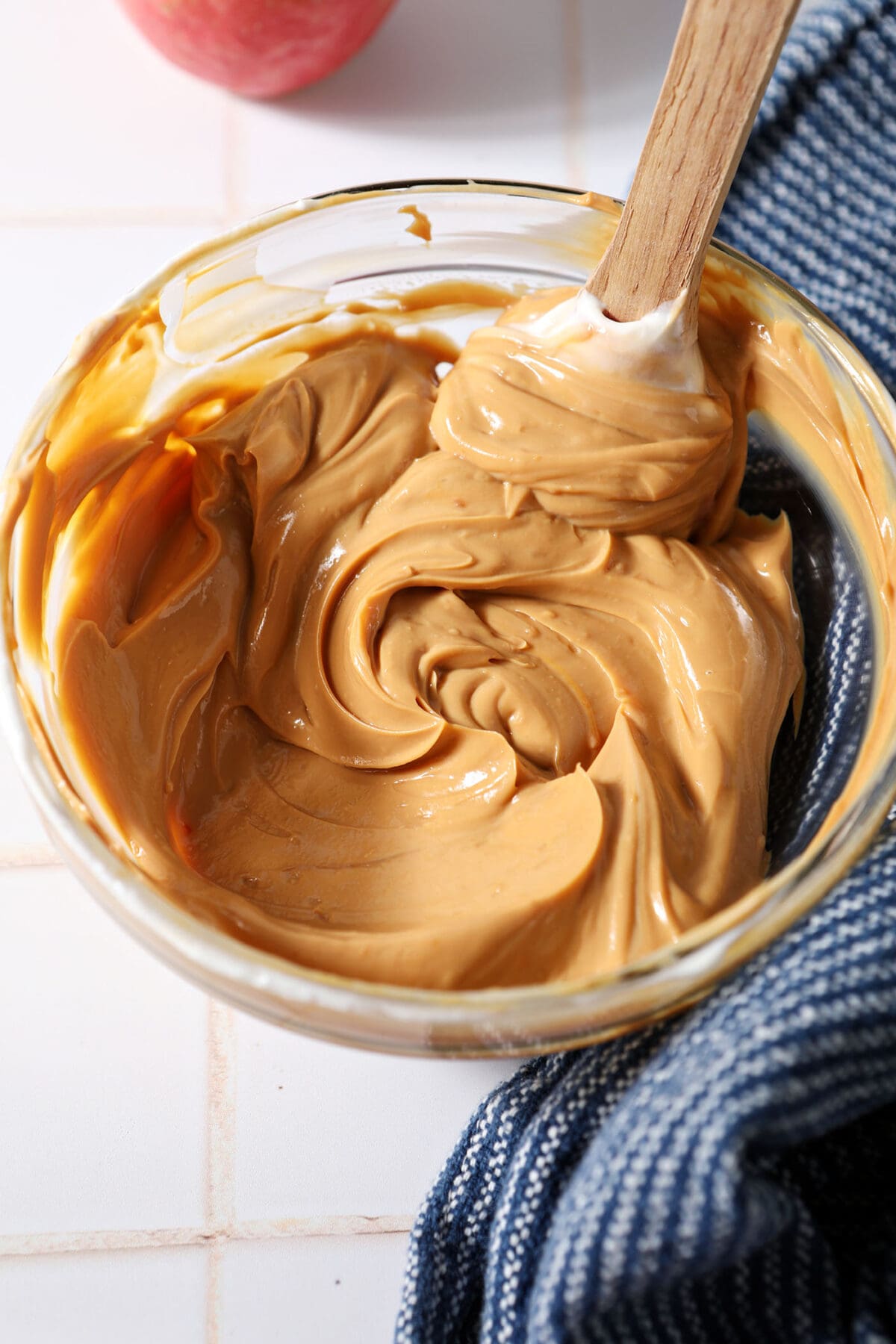 Mixed Dulce de Leche Caramel Dip in a bowl with a rubber spatula on a white tiled surface next to a blue linen