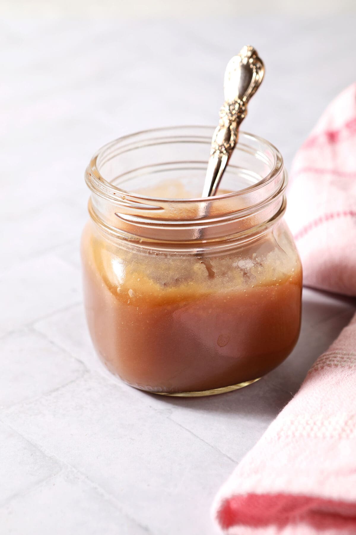 A mason jar of salted caramel sauce on a white tiled surface with a spoon inside it next to a pink linen