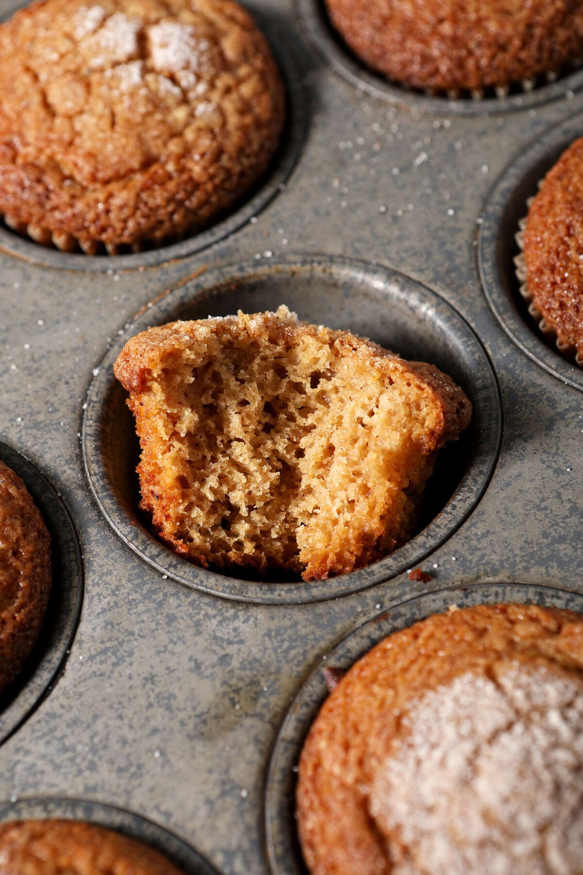 A bitten-into apple cider muffin sits in a tin next to more baked muffins