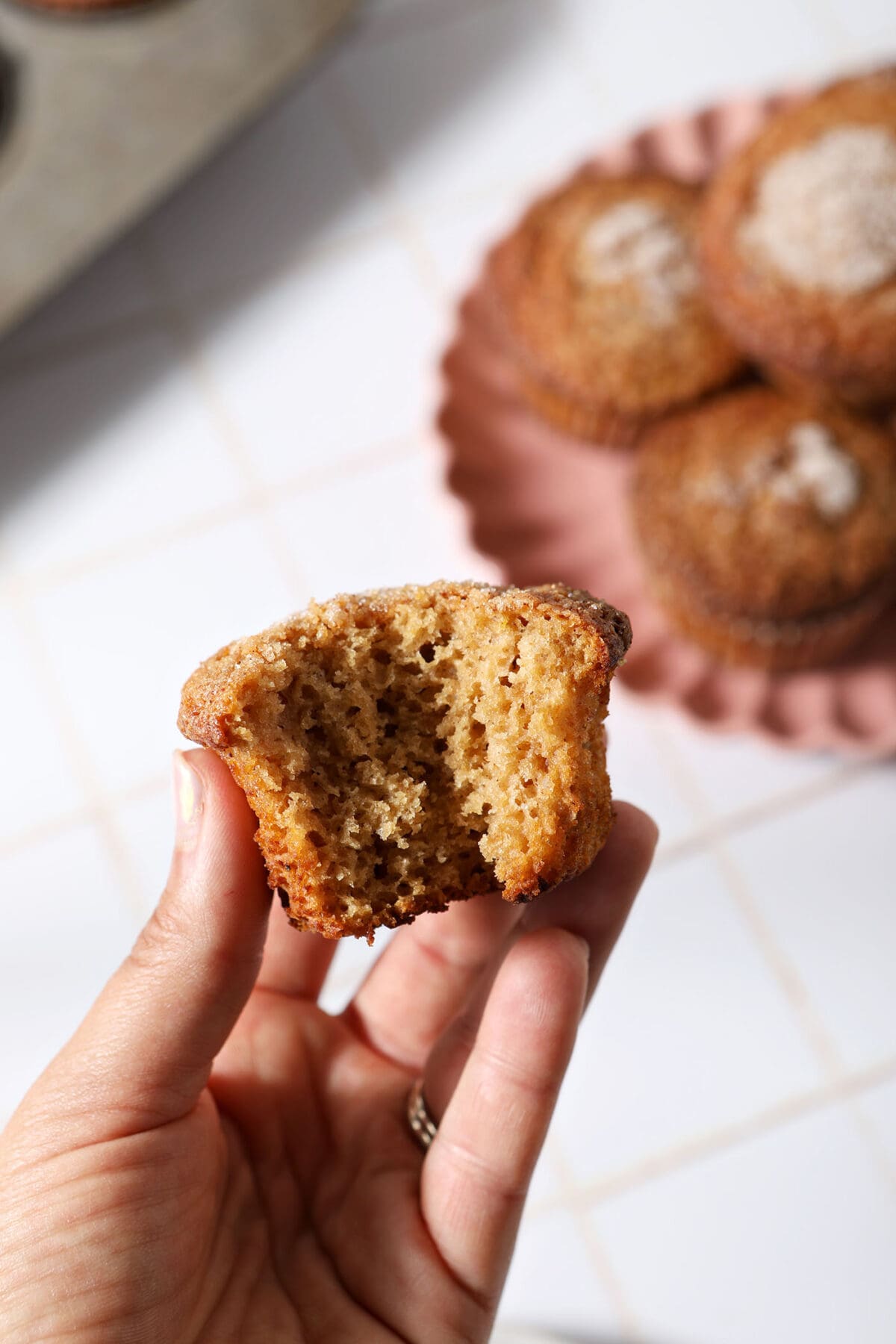A hand holds a bitten-into muffin above a pink plate of more muffins next to a muffin tin on a white tiled surface