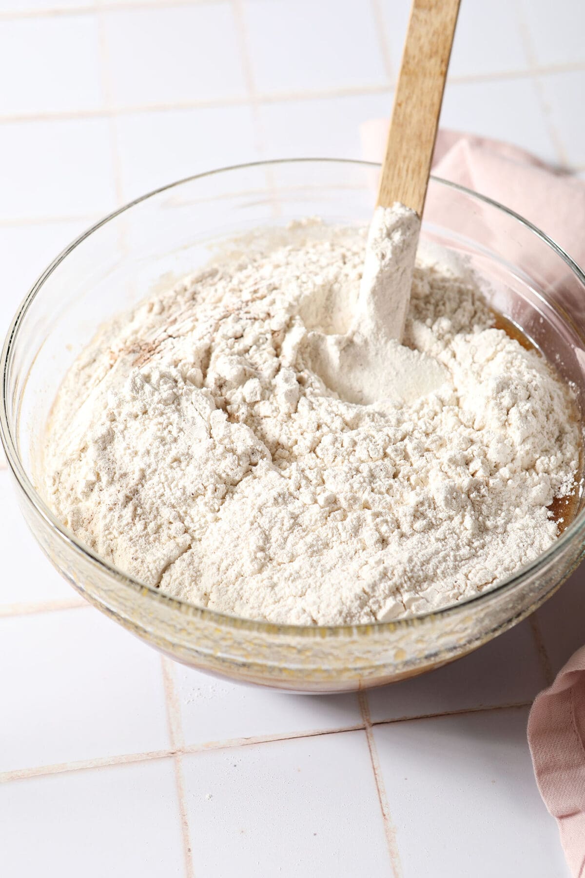 Dry ingredients on top of wet ingredients in a glass bowl before mixing on a white tiled surface next to a pink linen