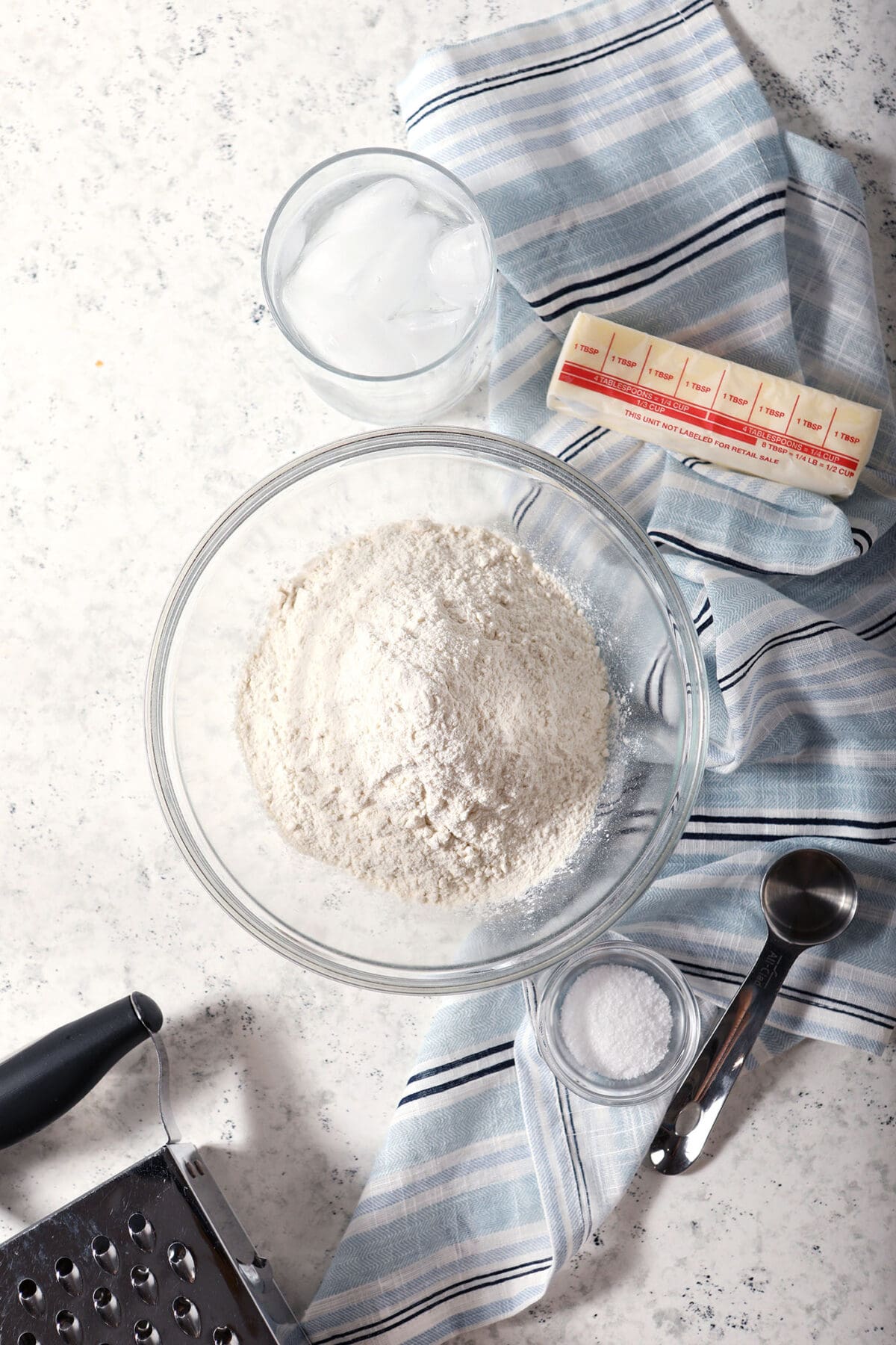 Ingredients to make pie crust on a white surface in bowls with a blue striped linen