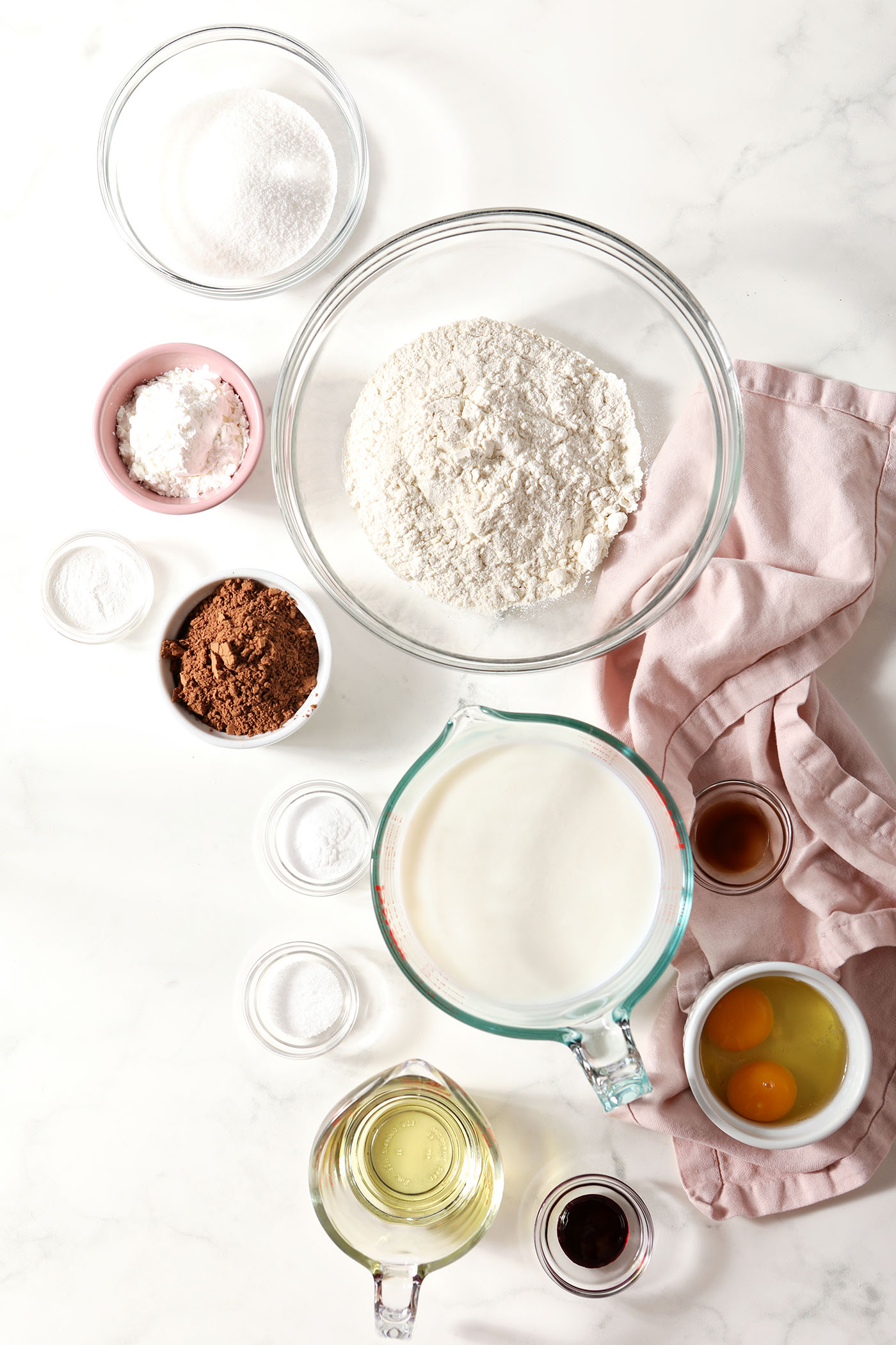 Ingredients to make waffles into red velvet in bowls and measuring cups on a white surface with a pink linen