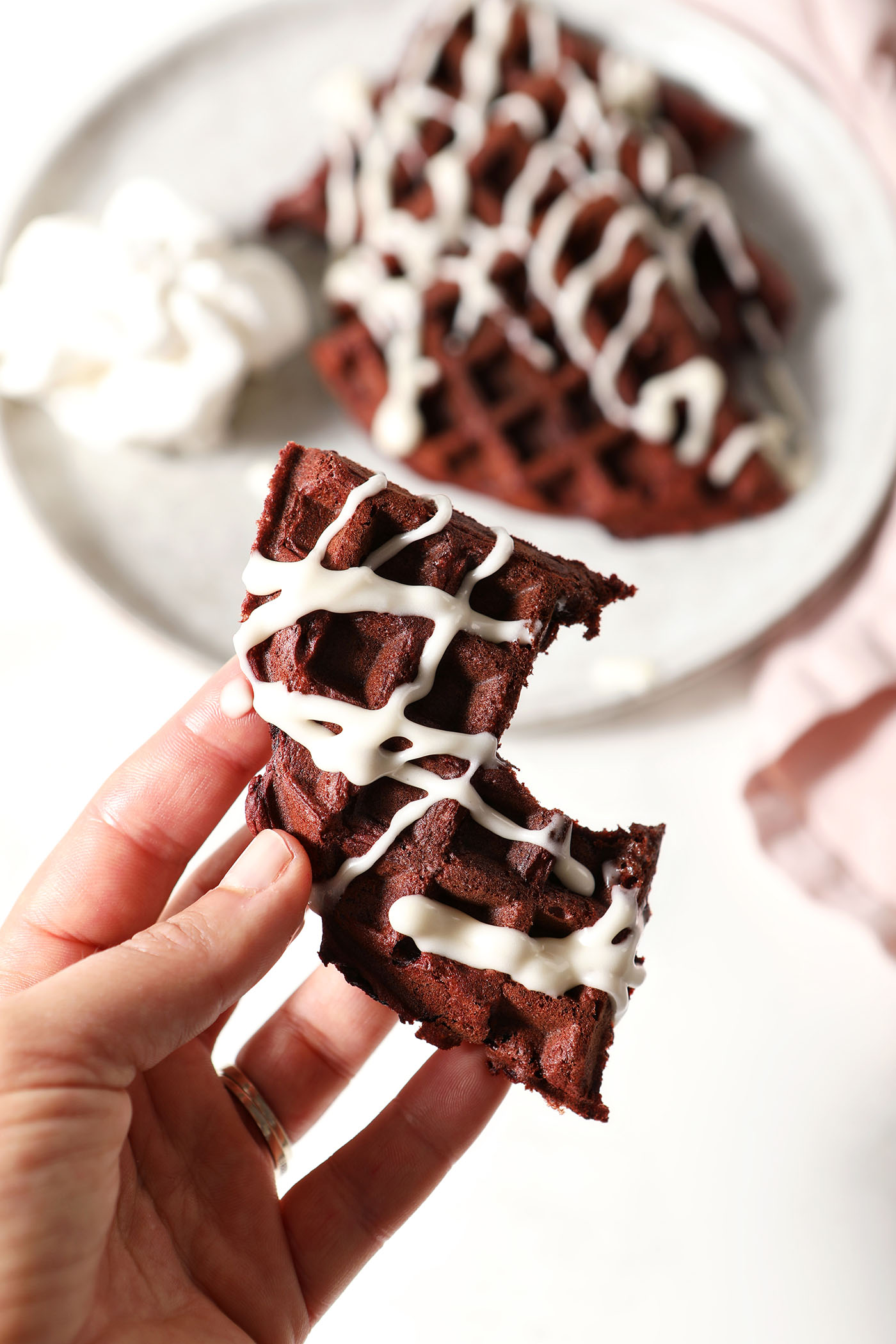 A hand holds a bitten-into red velvet waffle drizzled with cream cheese icing above a plate with more portions of waffles