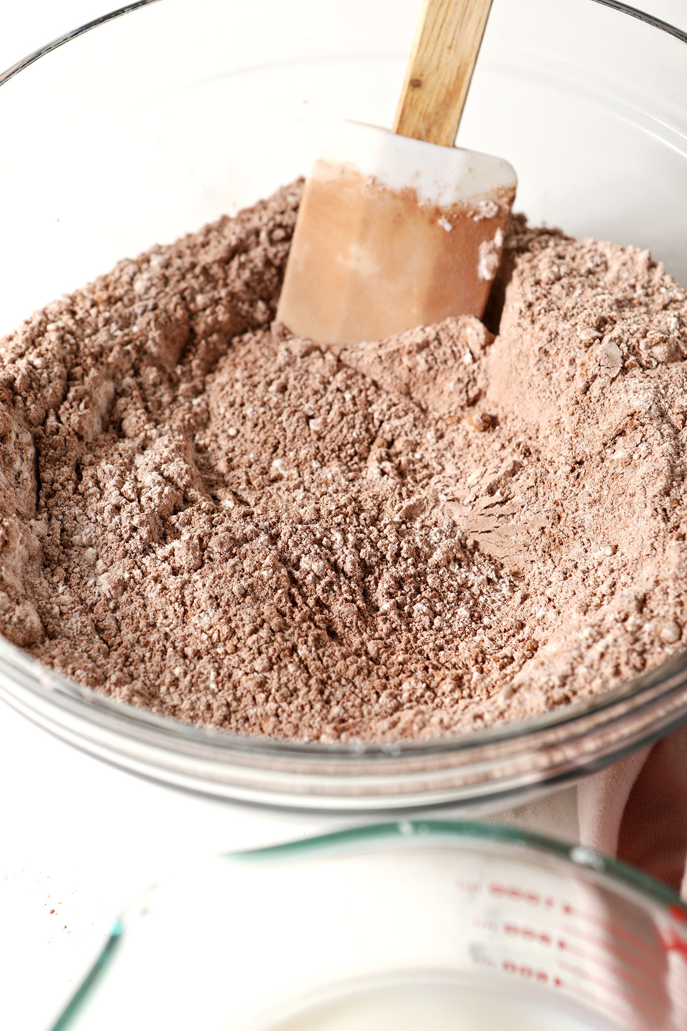 Cocoa powder and other dry ingredients in a glass bowl with a rubber spatula on a white surface