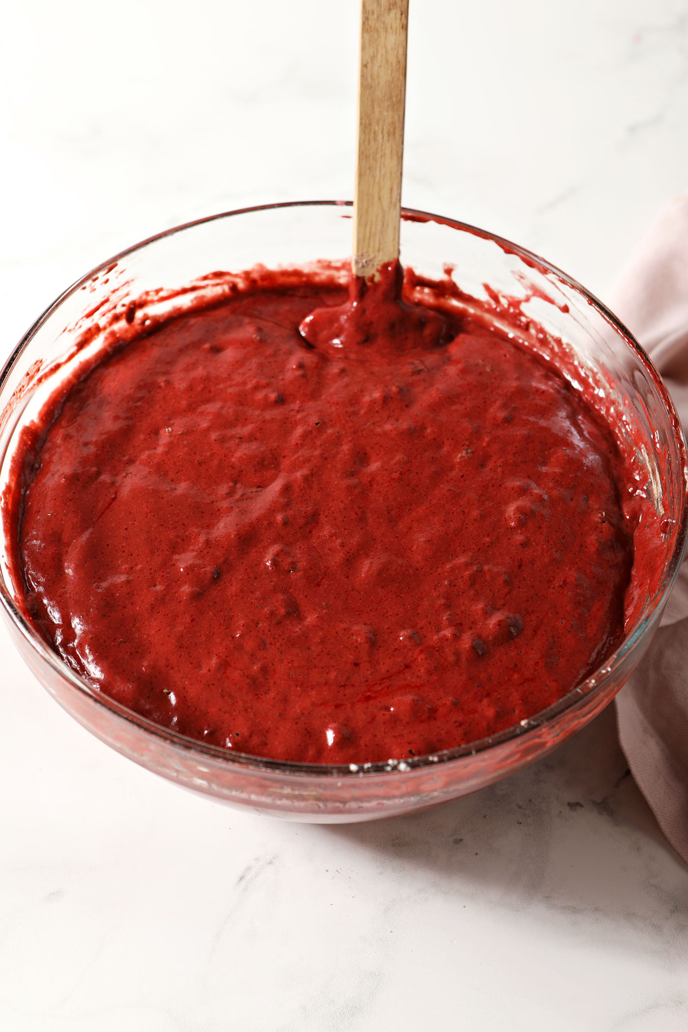 Red velvet waffle batter in a bowl with a rubber spatula on a white surface next to a pink linen