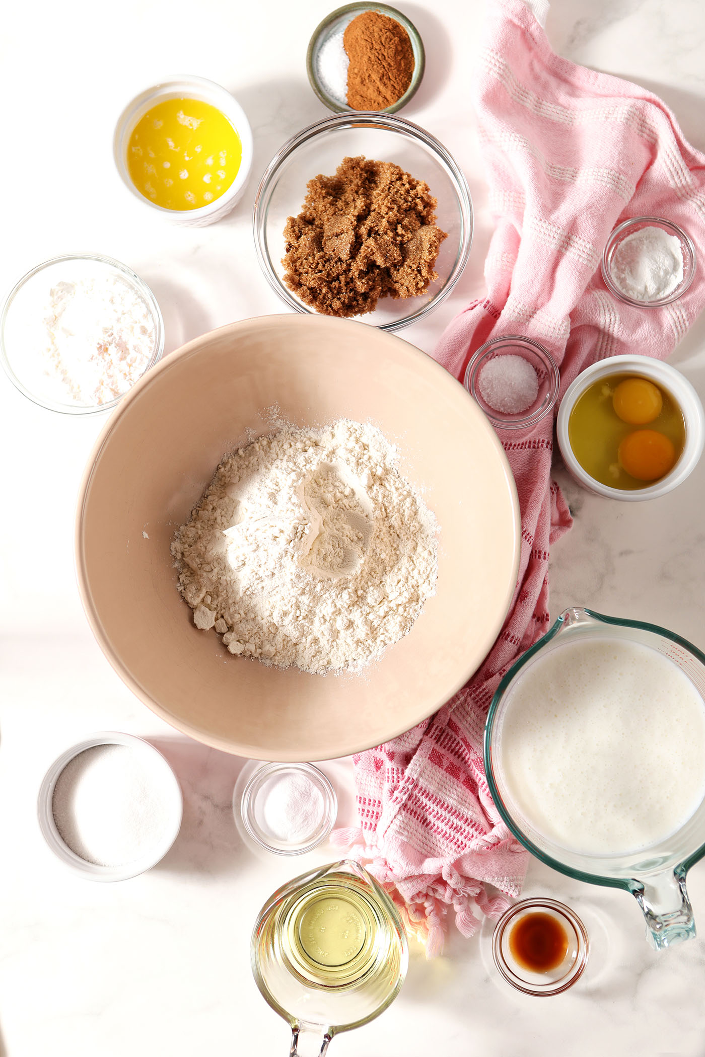 Ingredients to make homemade Cinnamon Roll Waffles in bowls and cups on a marble surface with a pink linen