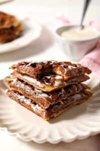 A stack of Cinnamon Roll Waffle quarters on a white scalloped plate on a marble surface with a pink linen