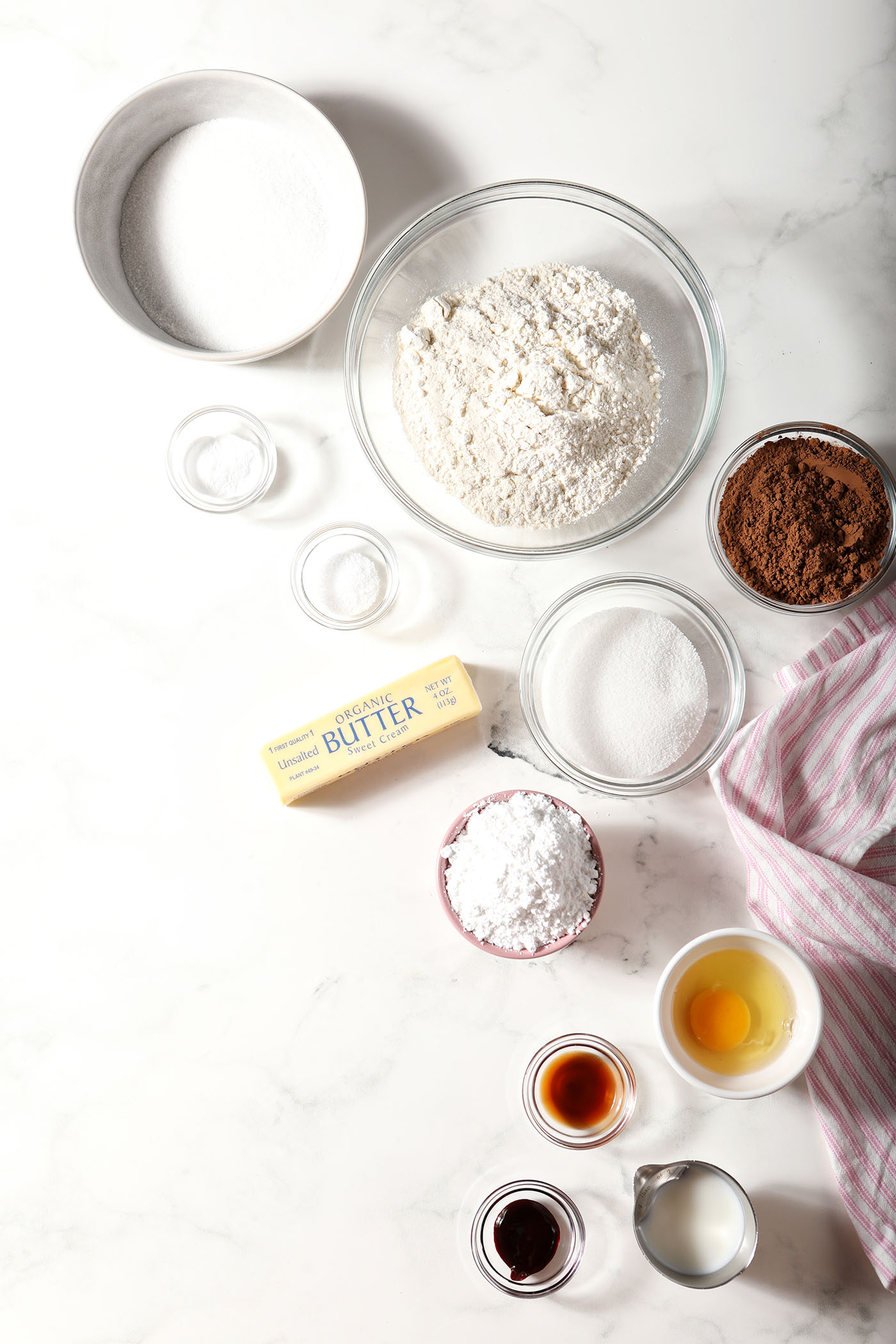 Ingredients to make red velvet sugar cookies in bowls on a marble surface with a pink and white striped linen