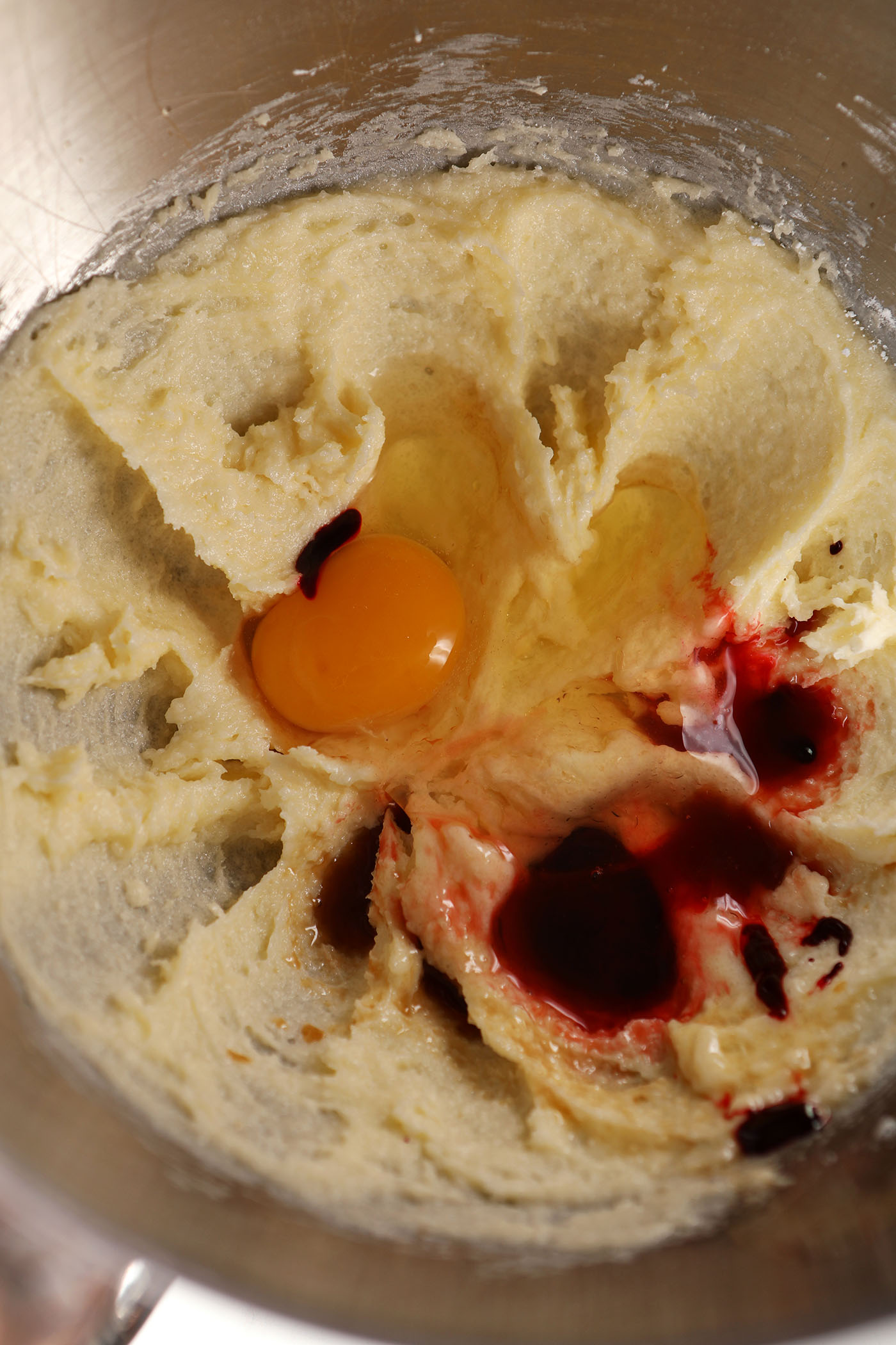 An egg, vanilla extract and red food coloring on top of creamed butter and sugar in a metal bowl