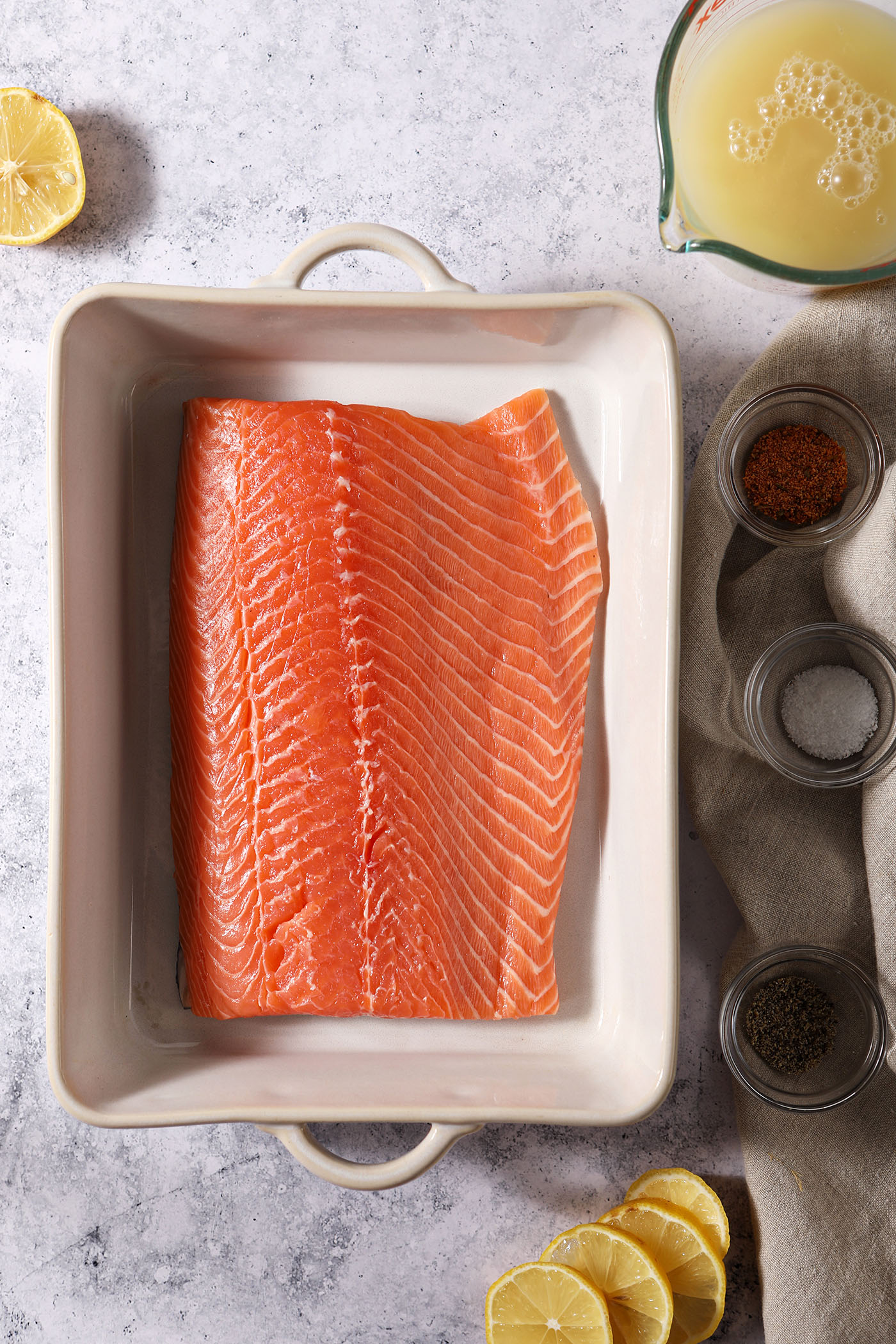 Overhead of a large cut of salmon in a white casserole dish surrounded by spices, a bowl of liquid and lemon wheels