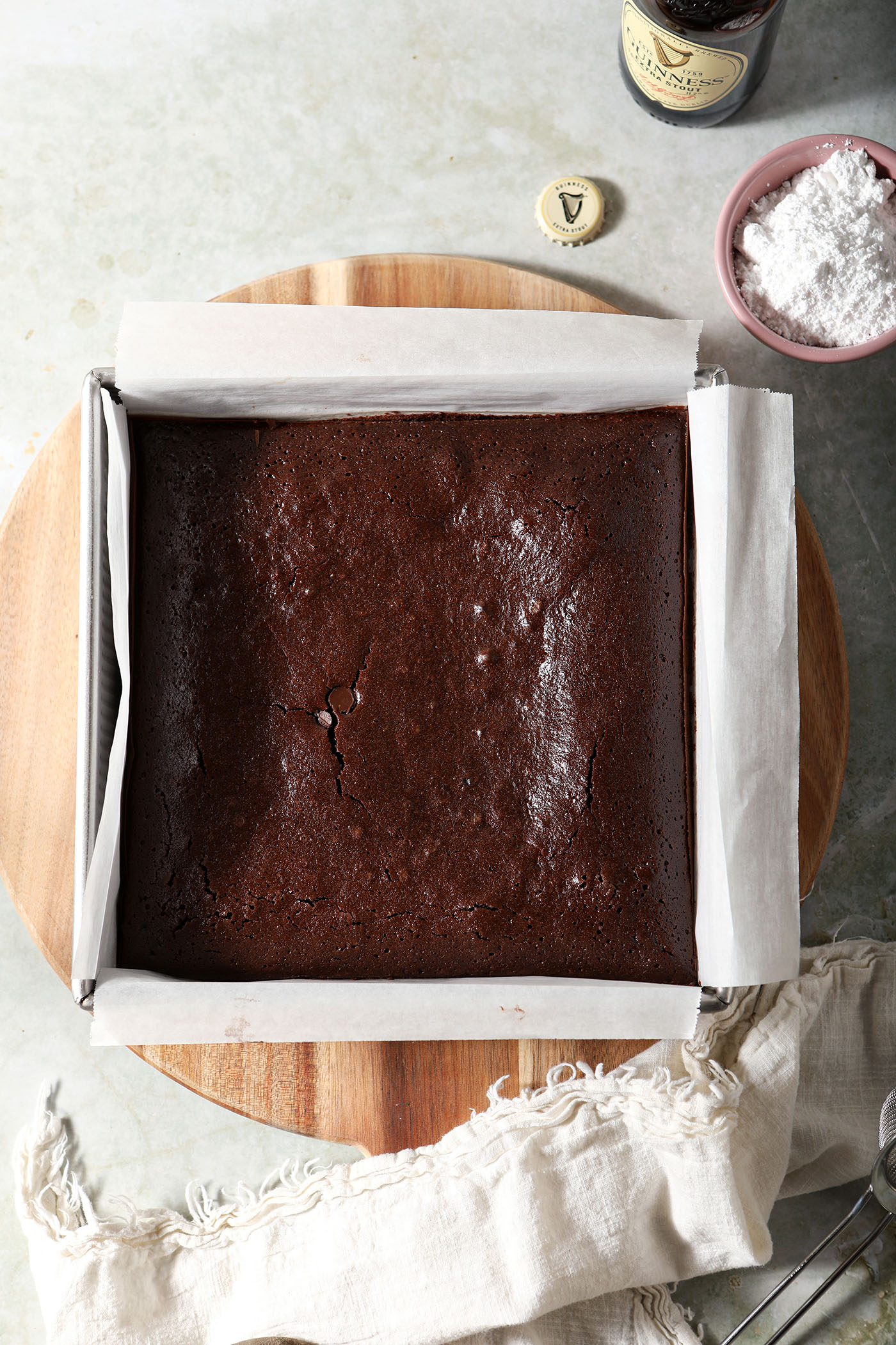 Baked Guinness brownies in a tin on top of a wooden board on a green countertop next to a bowl of powdered sugar and beer bottle