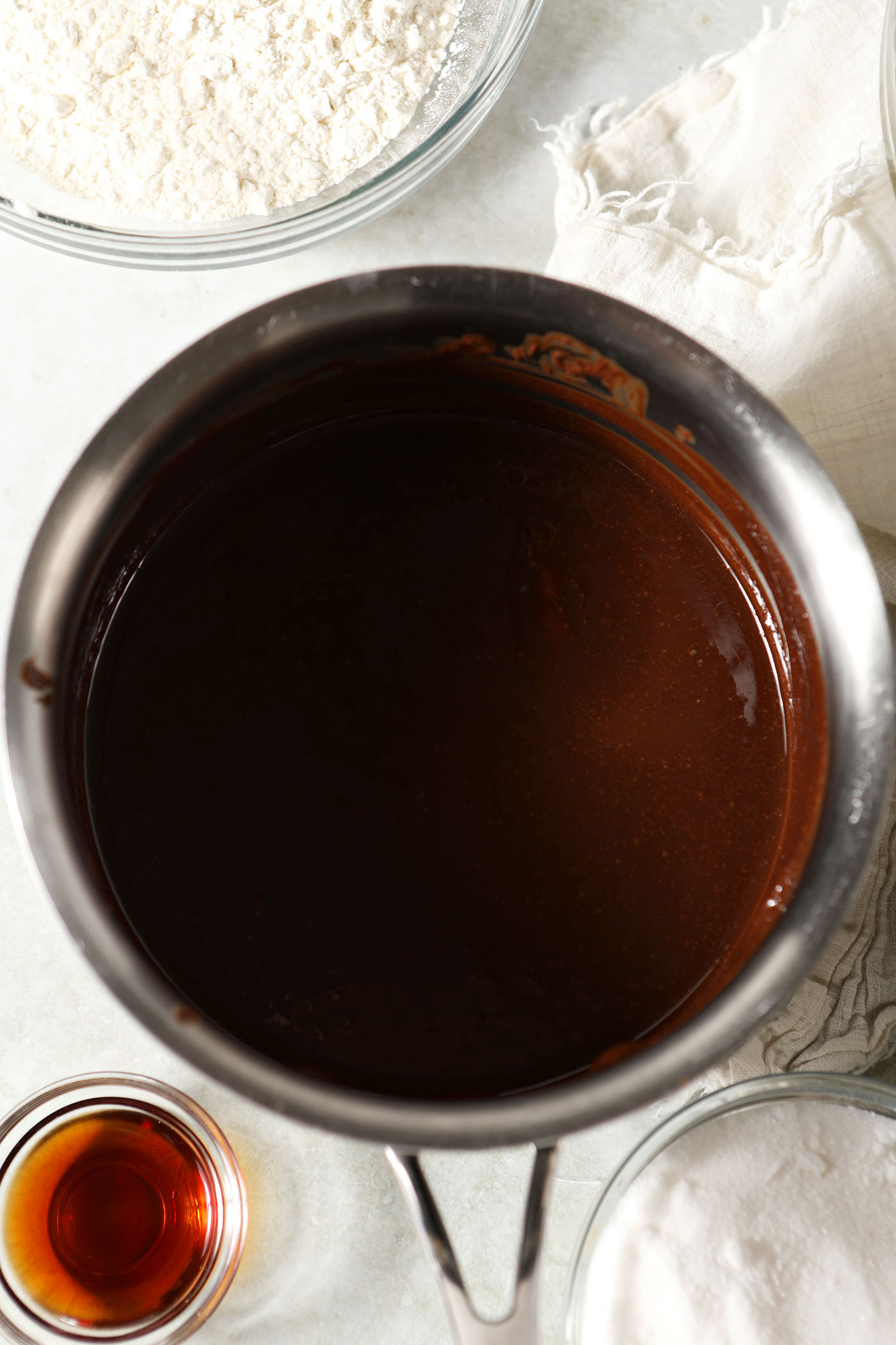 Melted chocolate and butter in a saucepan on a green countertop surrounded by bowls of other ingredients