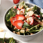 Close up of a bowl of strawberry salad with spinach, apples, nuts and feta on marble next to a blue linen