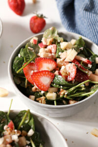 Close up of a bowl of strawberry salad with spinach, apples, nuts and feta on marble next to a blue linen