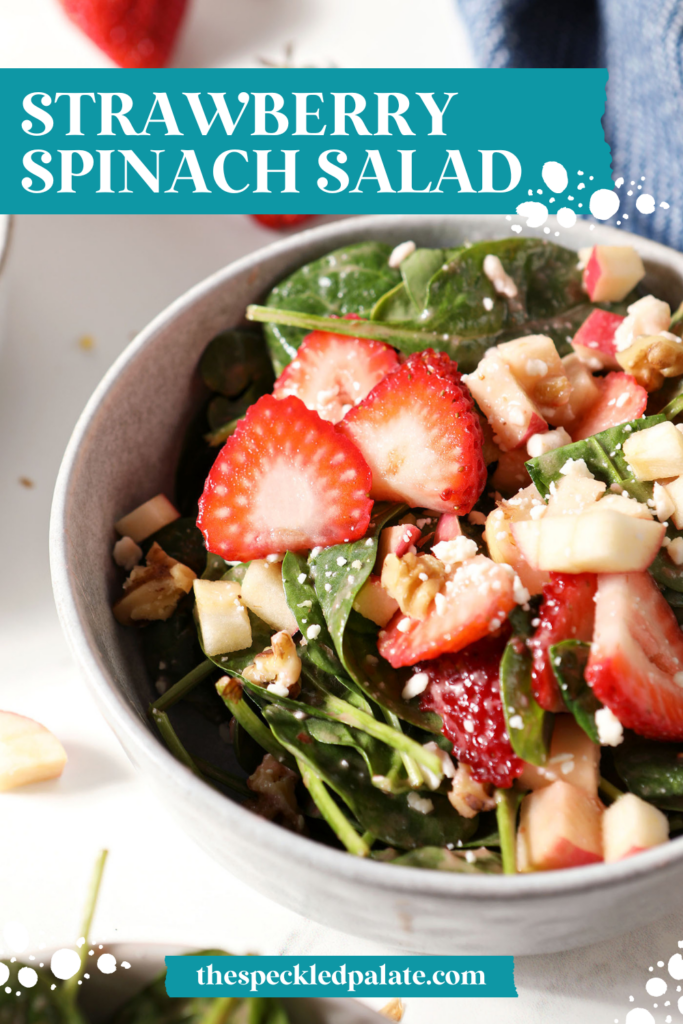 A bowl of strawberry salad with spinach, apples, nuts and feta on marble next to a blue linen with the text strawberry spinach salad