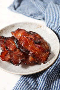 Close up of a small grey plate holding several slices of maple bacon next to a blue linen