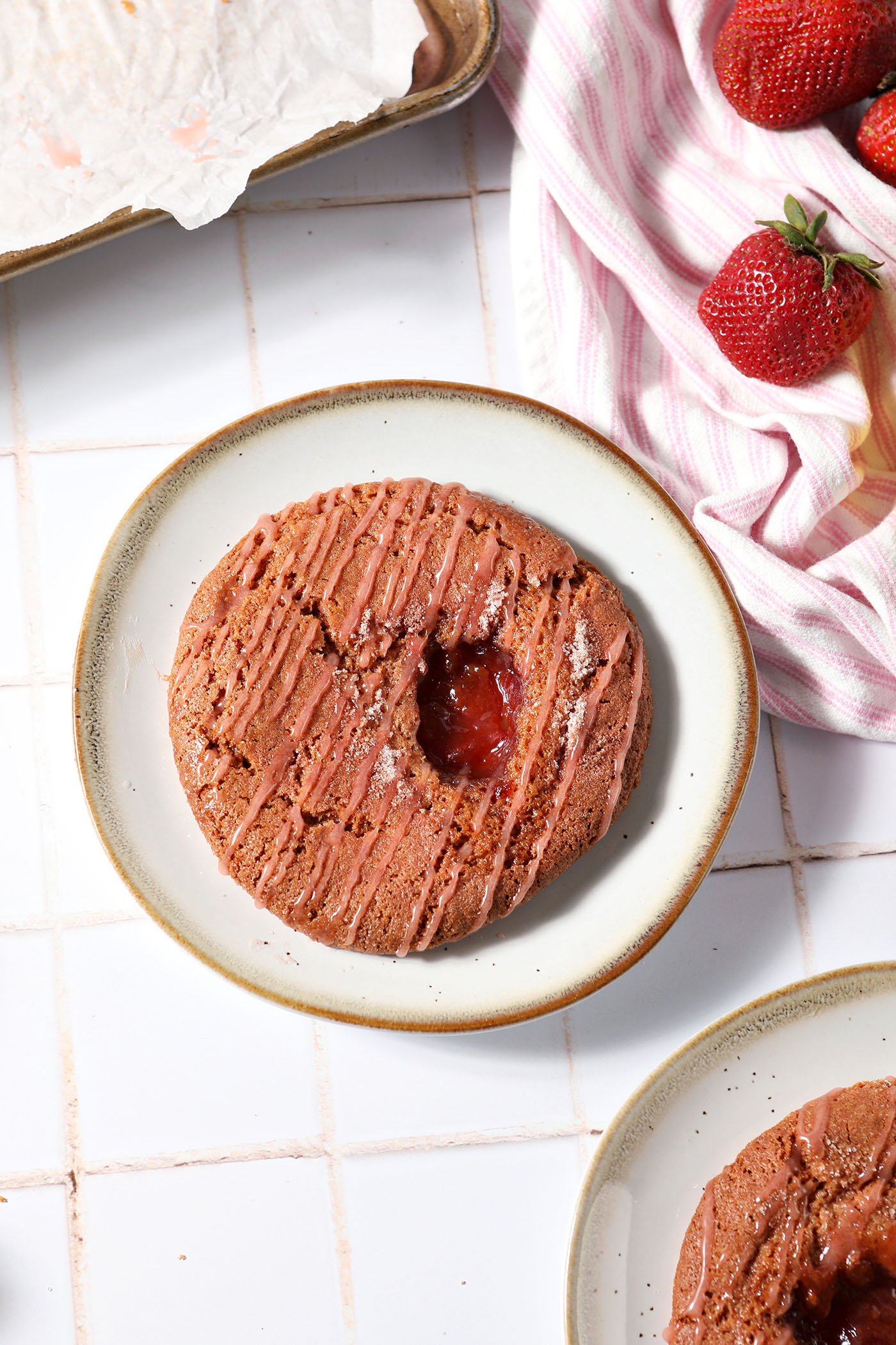 Two Strawberry Lemonade Cookies on plates on a tiled white surface with a pink and white striped linen and fresh strawberries